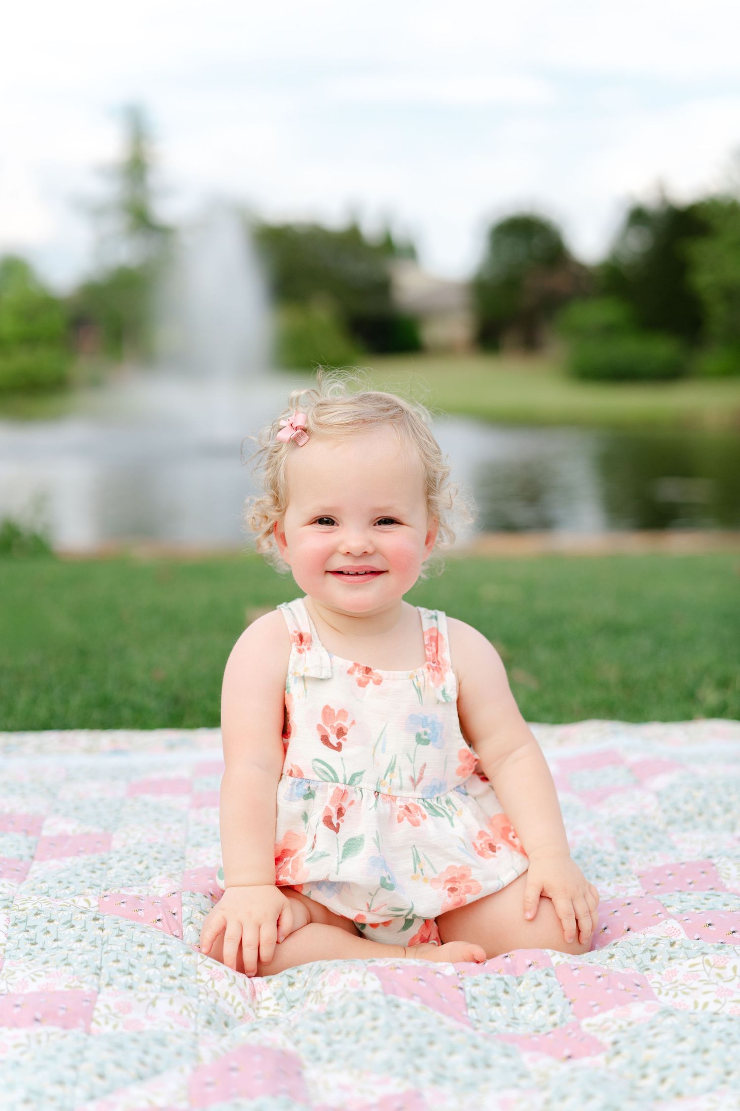 Smiling toddler girl in floral romper sitting on a patchwork quilt outdoors near a pond with a fountain.