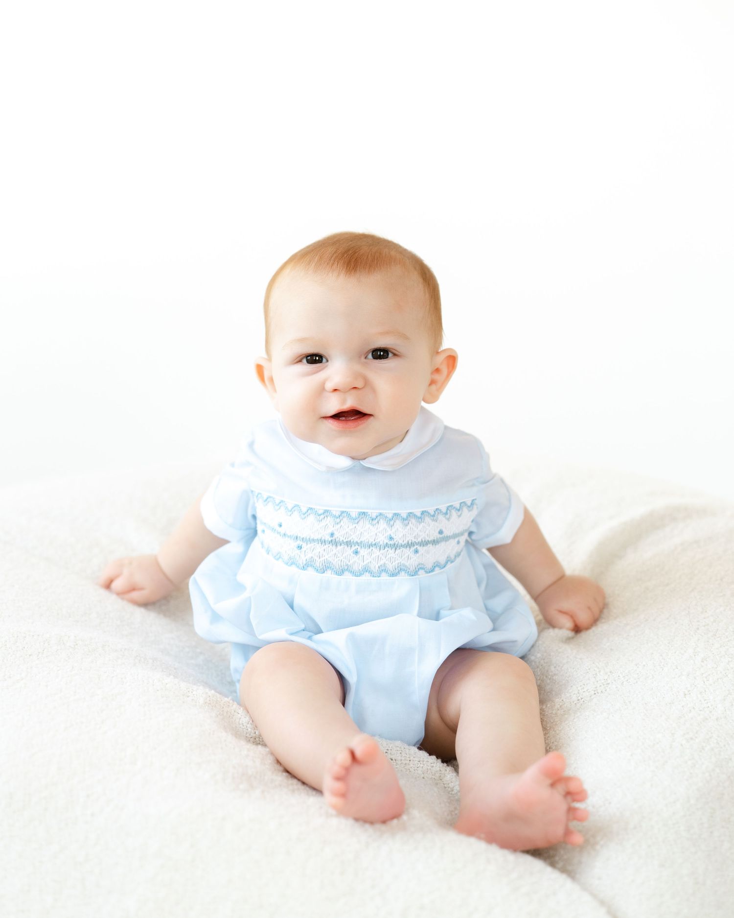 Baby in blue smocked romper sitting on white blanket, smiling happily in a professional portrait photo.