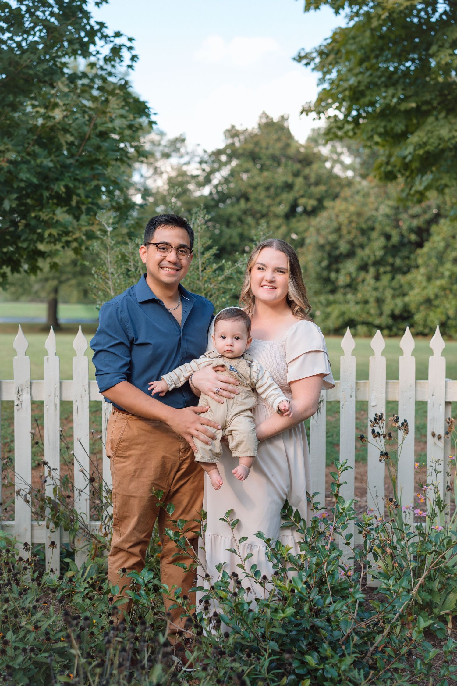 A young family with a baby poses in front of a white picket fence surrounded by lush greenery.