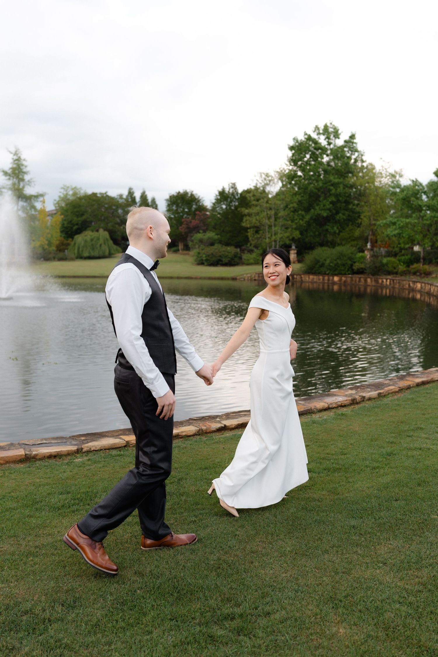 Bride and groom walk hand-in-hand by a pond with a fountain, surrounded by lush greenery on their wedding day.