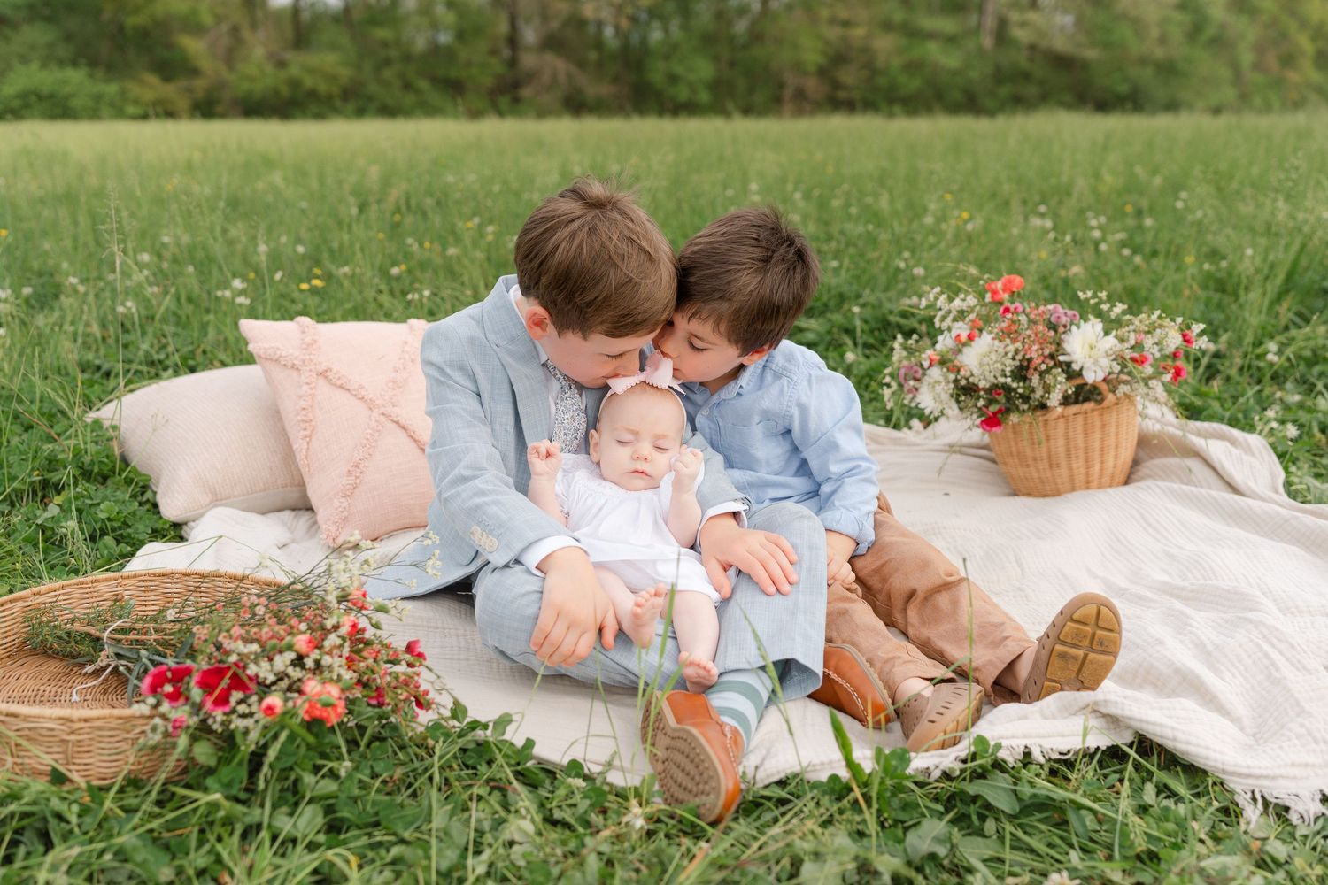 Two young brothers lovingly kiss their newborn sibling on a floral picnic blanket outdoors.