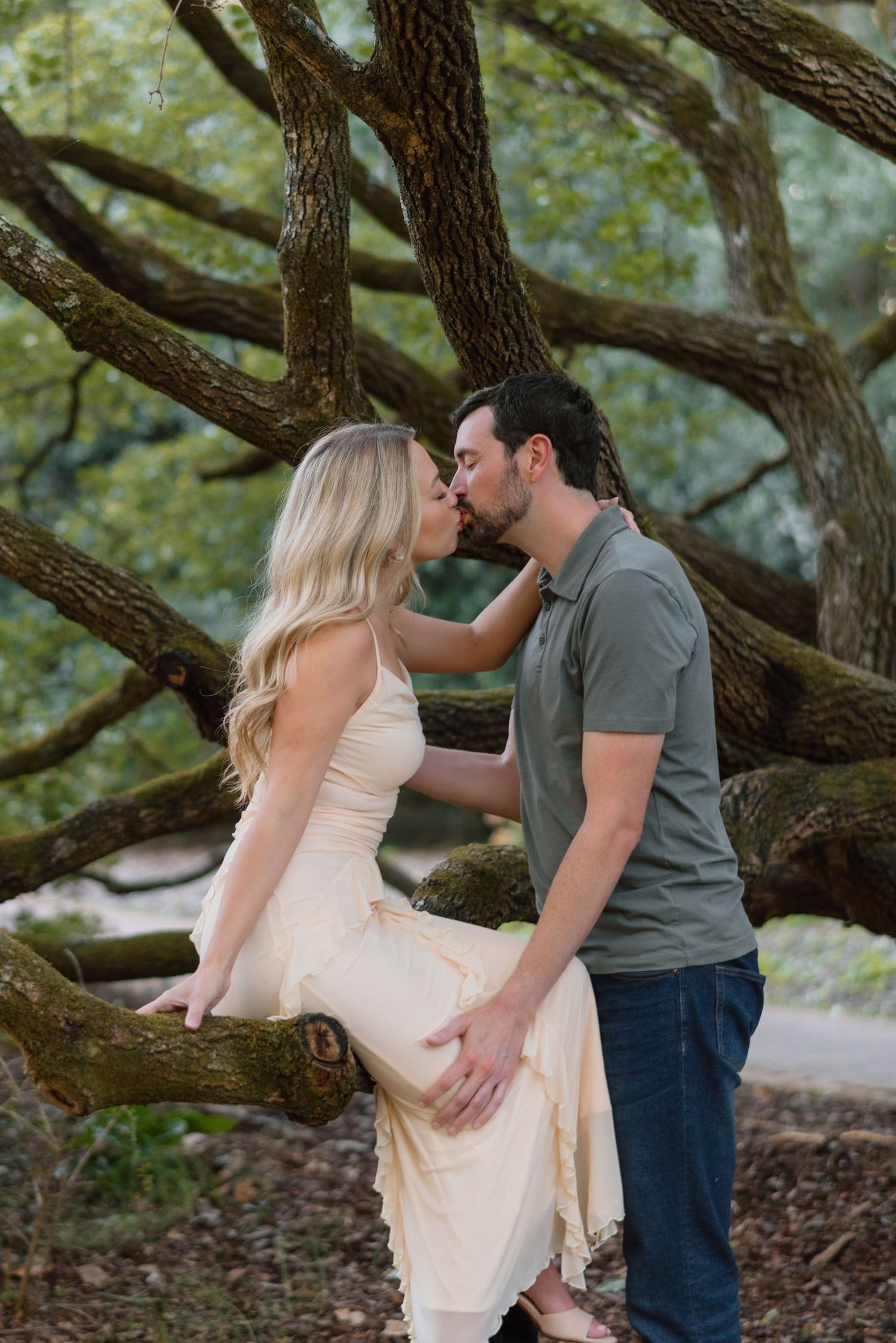 Couple kissing beside a massive oak tree, woman in cream halter dress seated on branch at golden hour.
