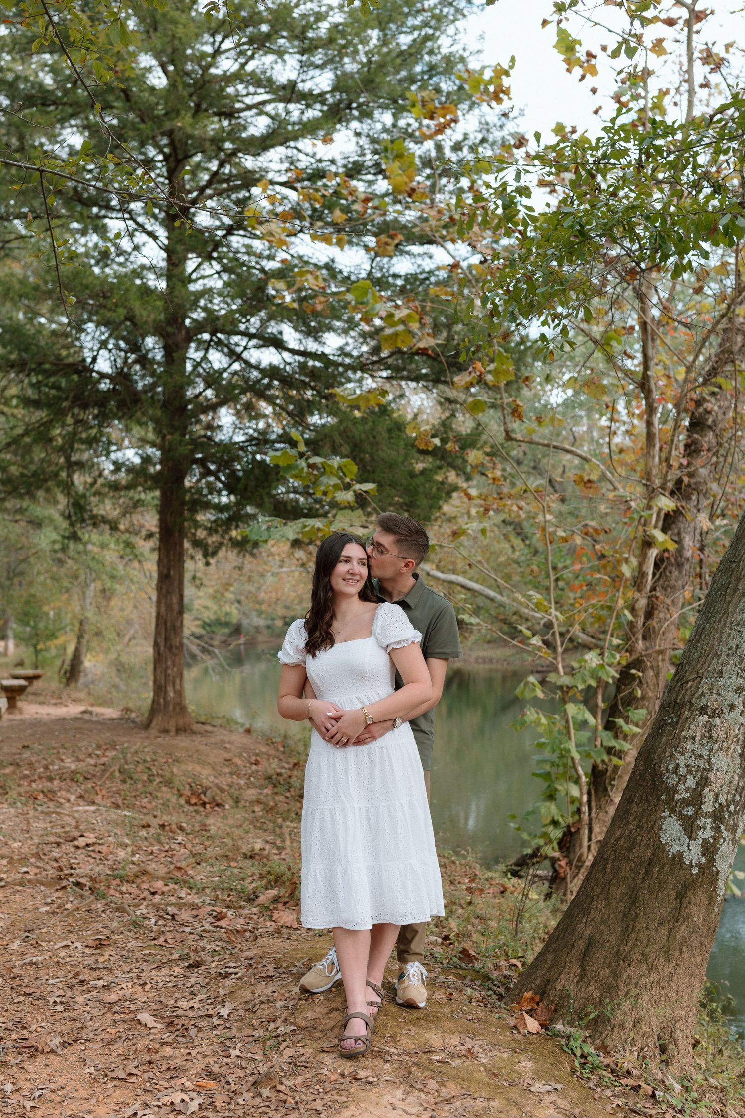 Man embracing woman in white midi dress near a serene woodland lake surrounded by tall pine trees.