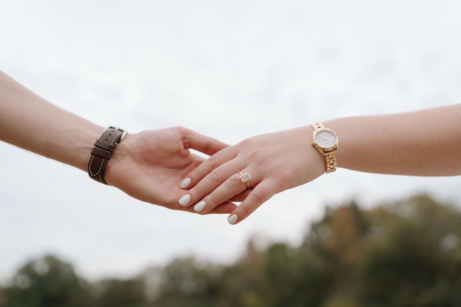 Close-up of couple holding hands, woman wearing gold watch and engagement ring, man wearing leather bracelet.