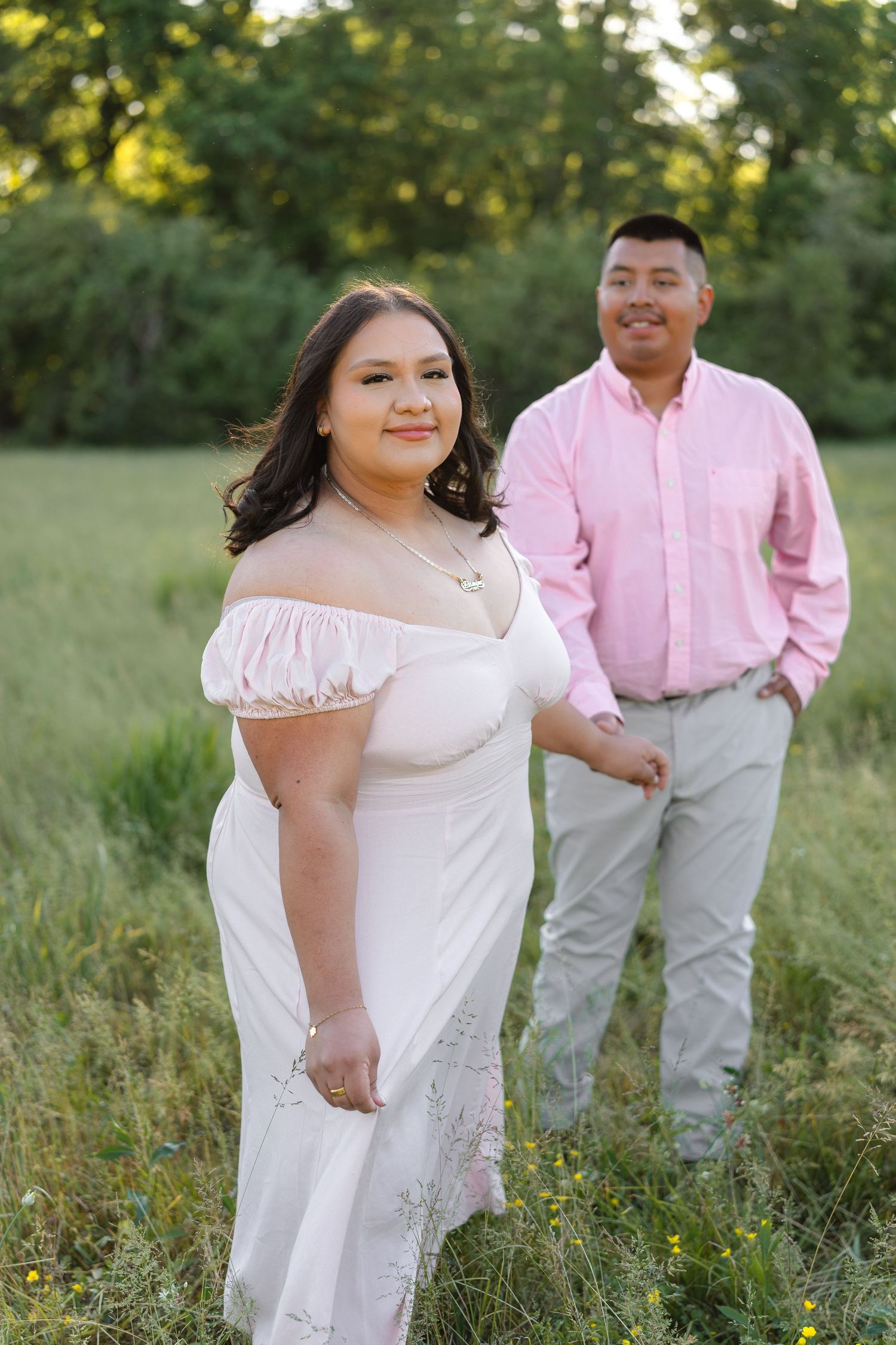 Couple in pink outfits posing in a green field, woman in white off-shoulder dress, man in pink shirt and gray pants.