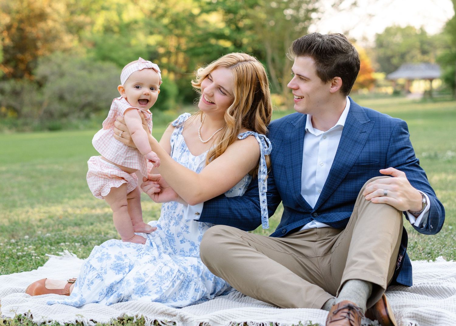 Young family sits on a picnic blanket in a park, laughing as baby girl stands on mom's lap.
