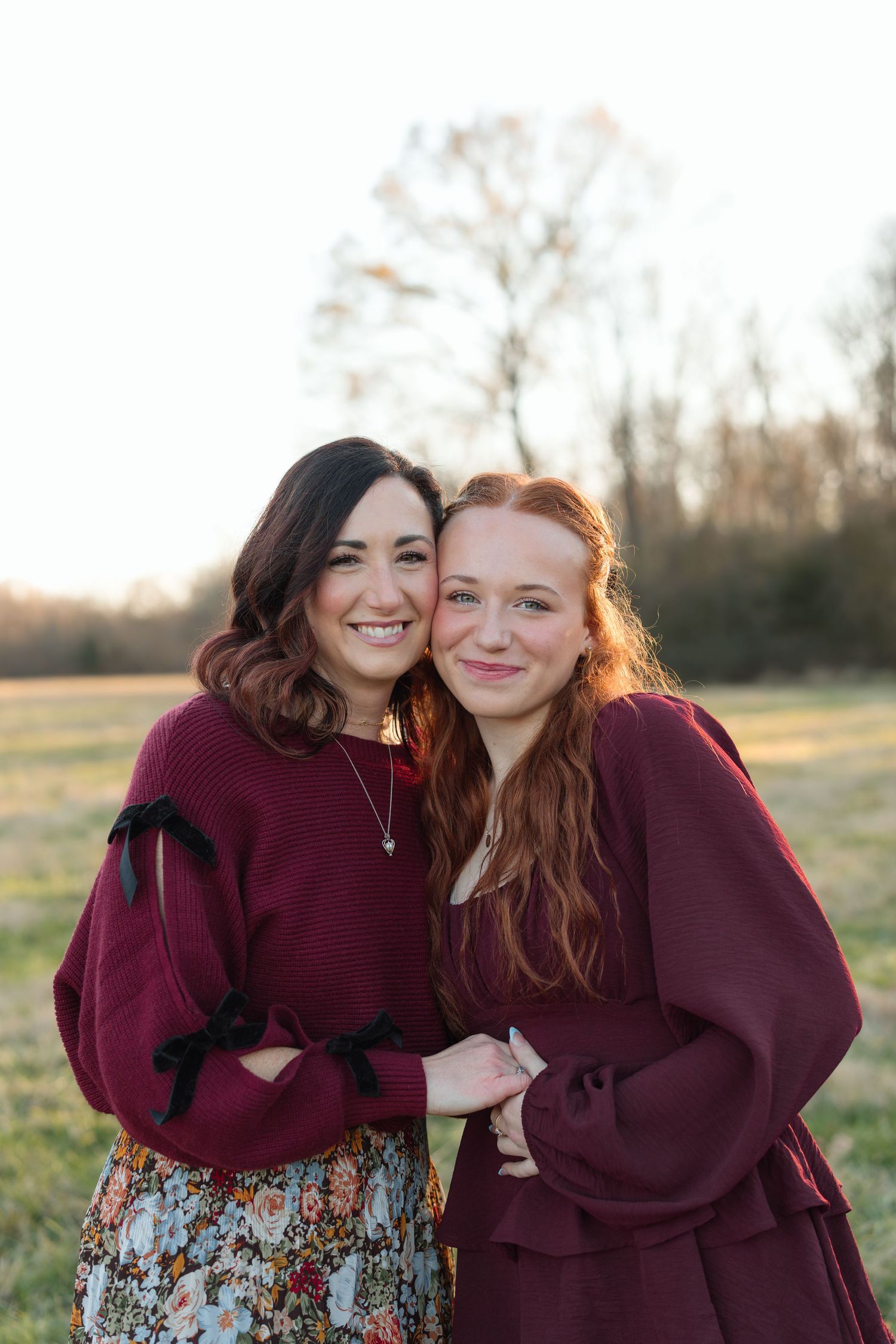 Two young women in burgundy outfits smiling together in a sunny outdoor field portrait session.