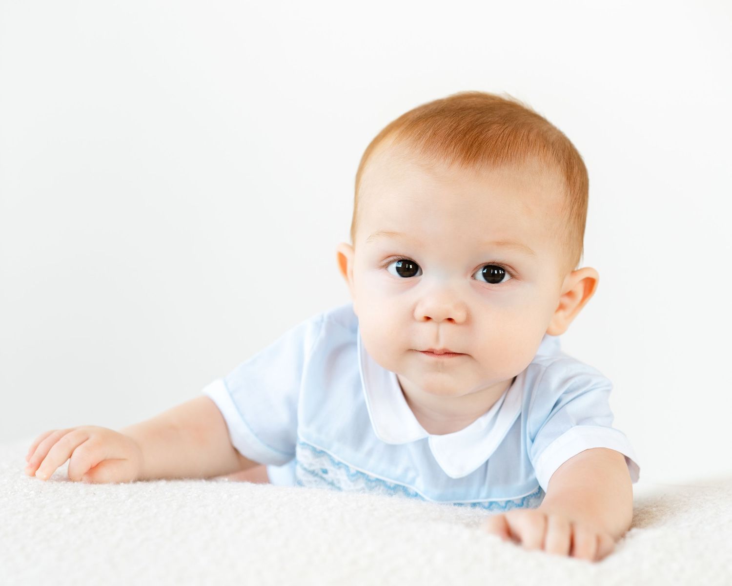 Adorable red-haired baby in a blue outfit doing tummy time on a soft white surface.