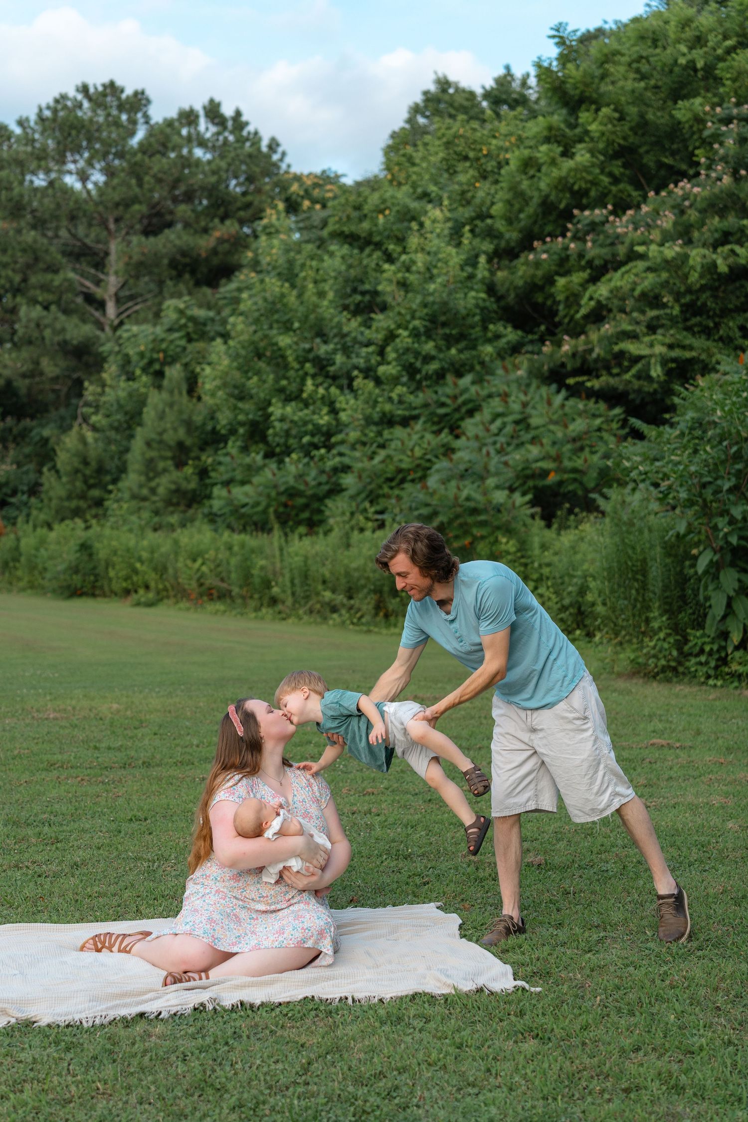 A happy family plays outdoors on a blanket in a lush green park, with parents playfully lifting their baby.