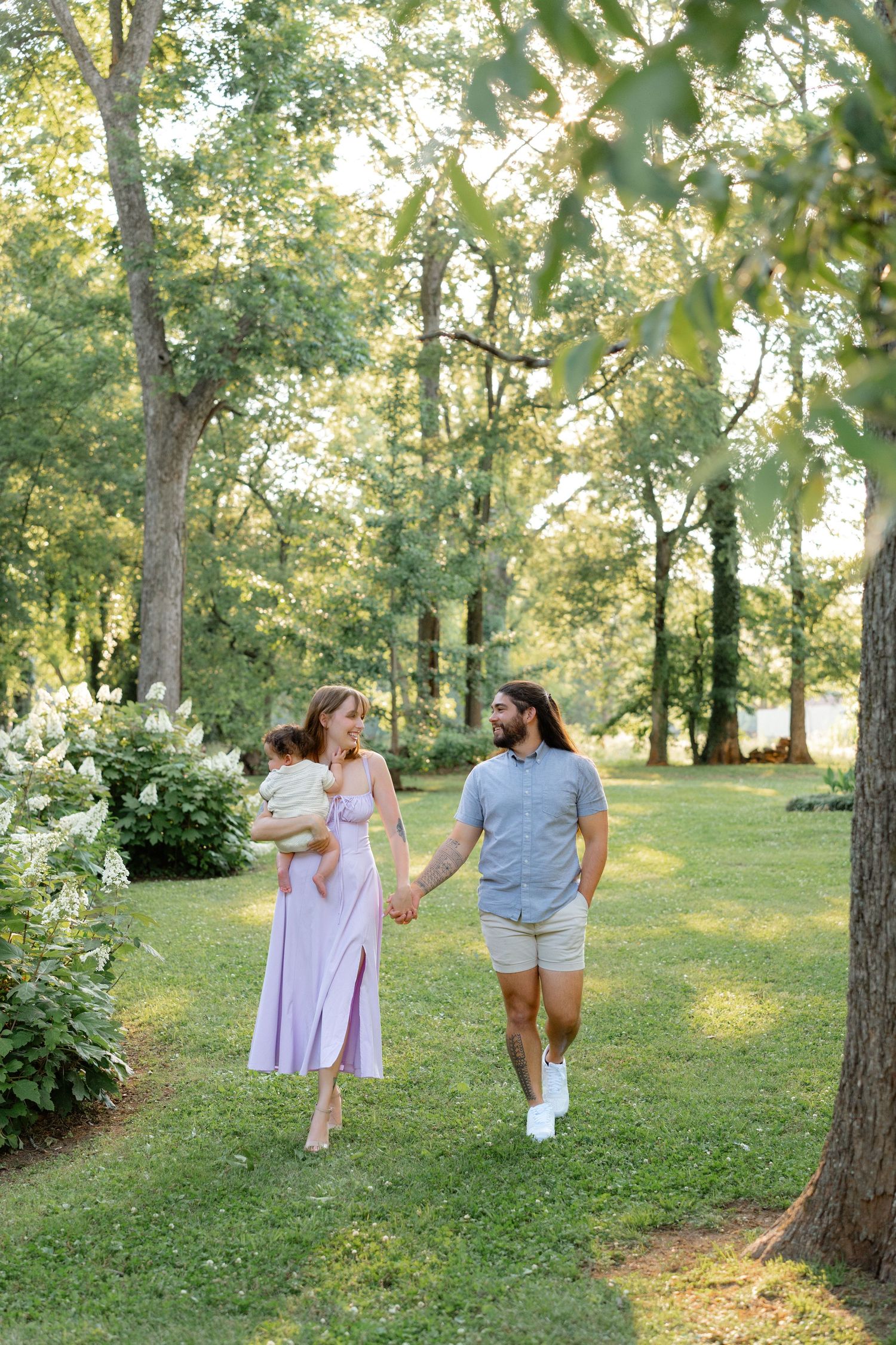 A young family walks hand-in-hand through a lush green park, with the mother holding a baby in a lavender dress.