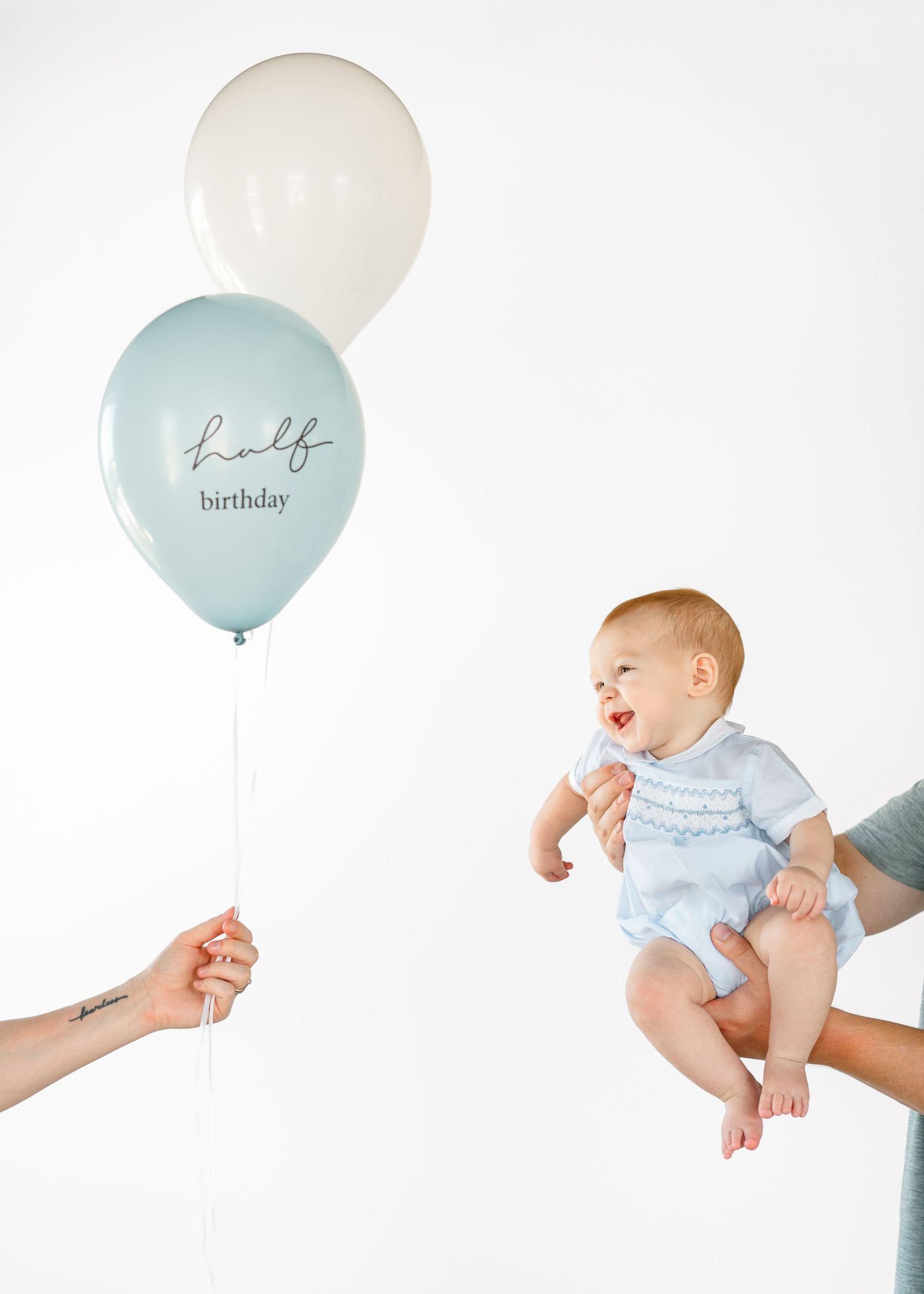 Baby in blue romper smiling at half birthday balloons held by adult hands on white background.