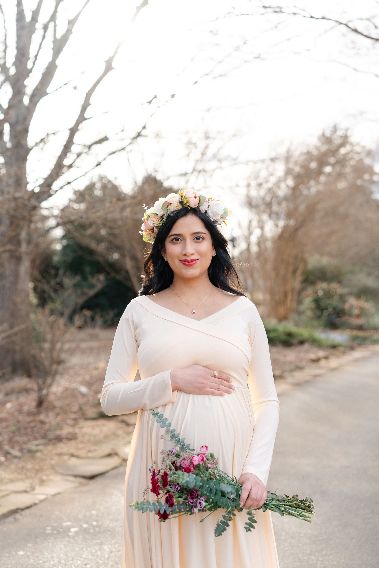 Pregnant woman in cream wrap maternity dress and floral crown holds bouquet outdoors.