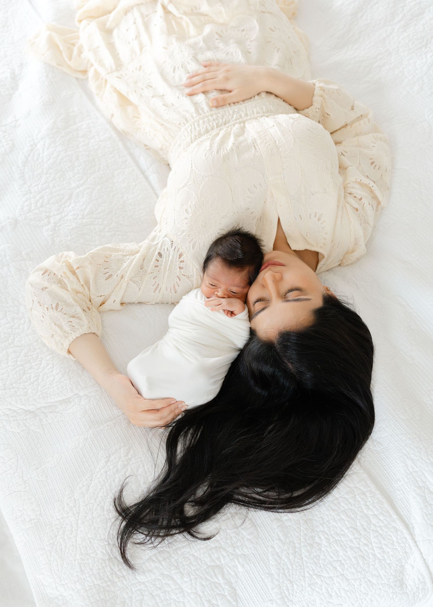 Aerial view of a dark-haired mother lying beside her swaddled newborn on a white bed.