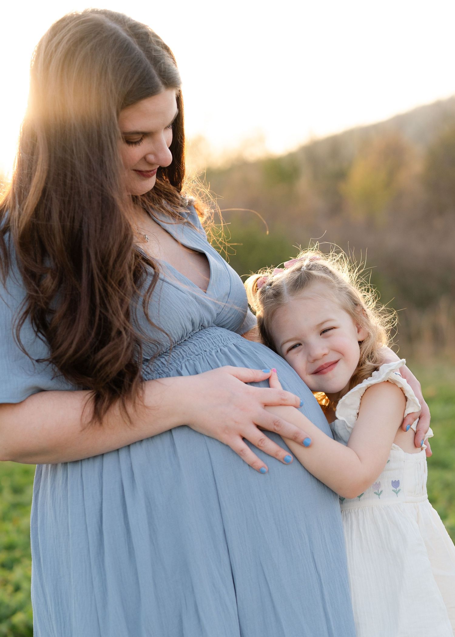 Pregnant woman in blue dress holds baby bump while toddler girl smiles and hugs her at golden hour.