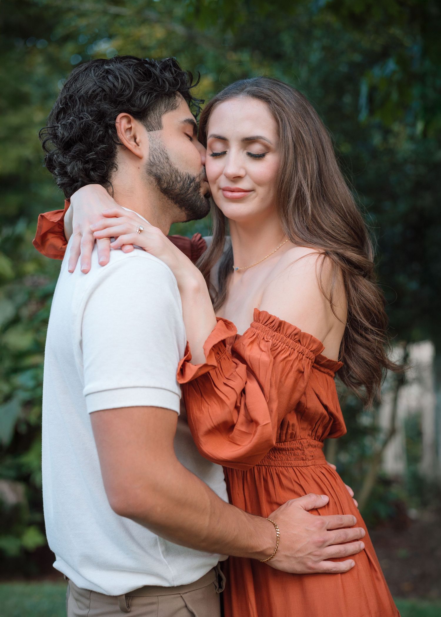 Romantic couple embracing outdoors, man kissing woman's cheek as she smiles in rust-colored off-shoulder dress.