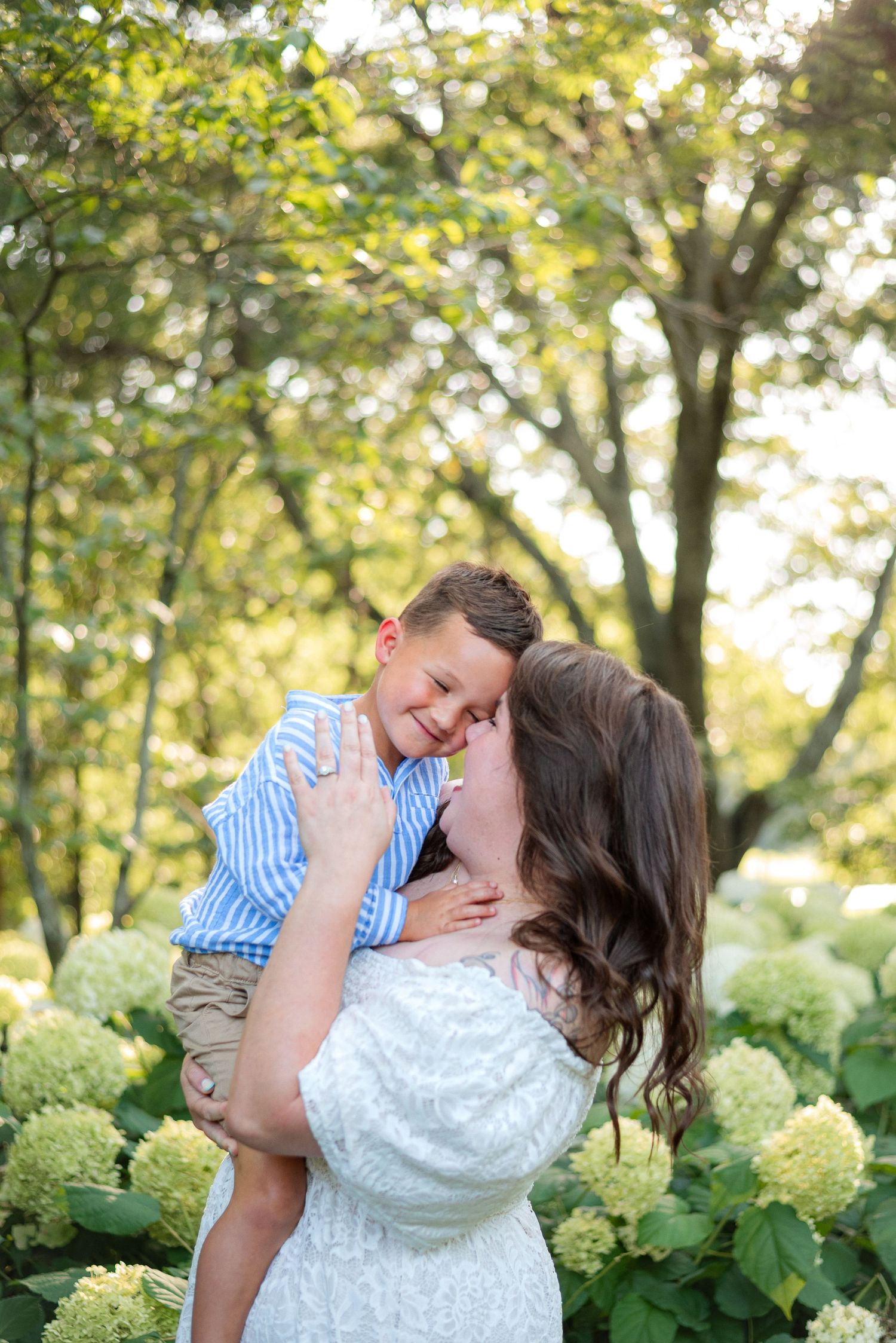Mother kisses smiling young son held in her arms among white hydrangea blooms outdoors.