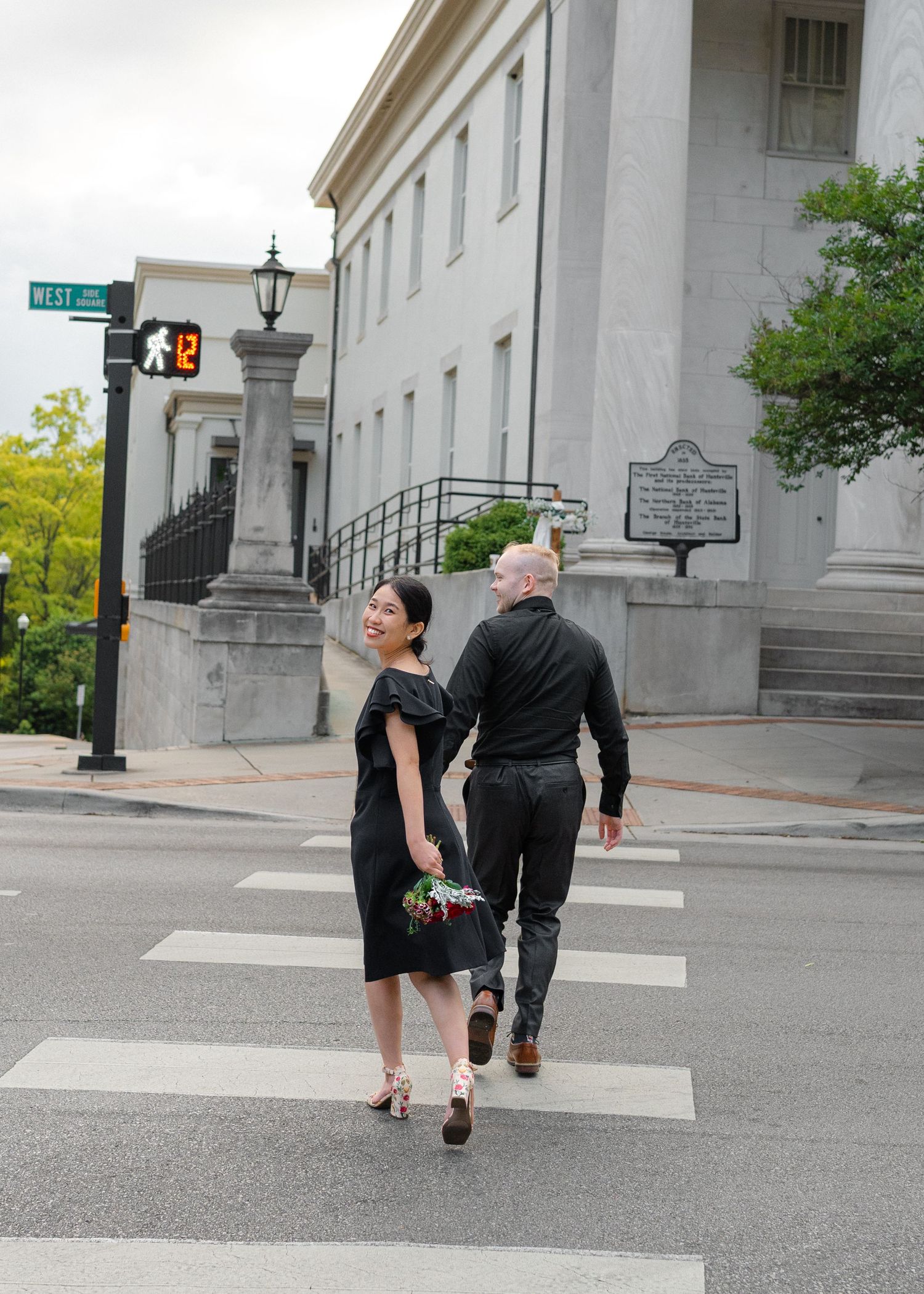 Couple in matching black outfits cross a street holding hands near a historic courthouse building.