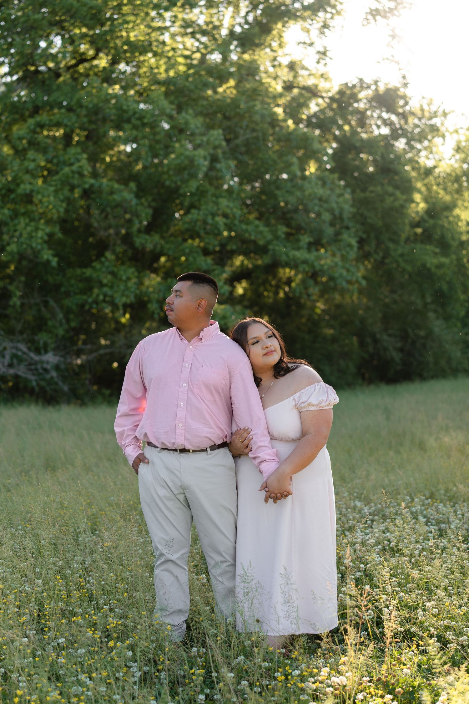 Couple posing in a sunny green meadow, man in pink shirt and woman in white off-shoulder dress, holding hands.