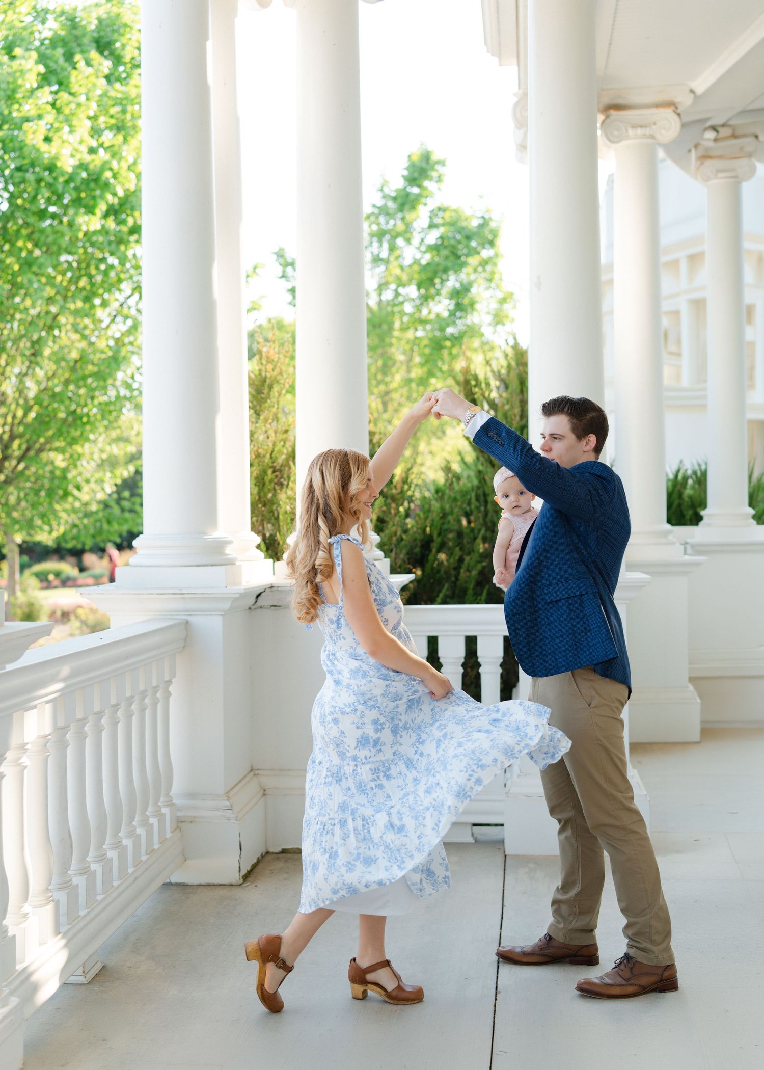 Couple dances on a white columned porch, man holding baby while twirling woman in floral dress.