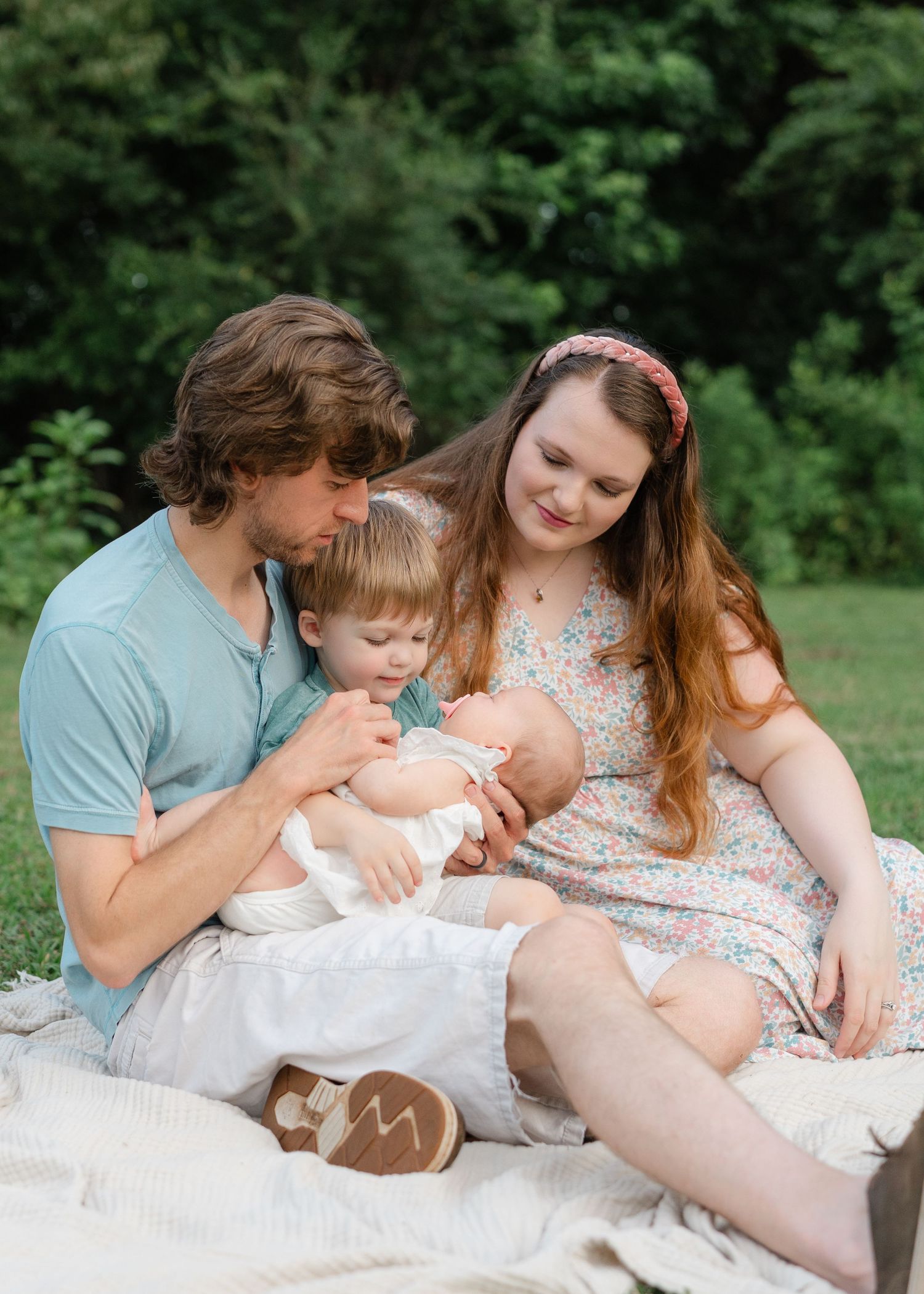Young family sitting on blanket outdoors, parents lovingly gazing at newborn baby with toddler nearby, lush green background.