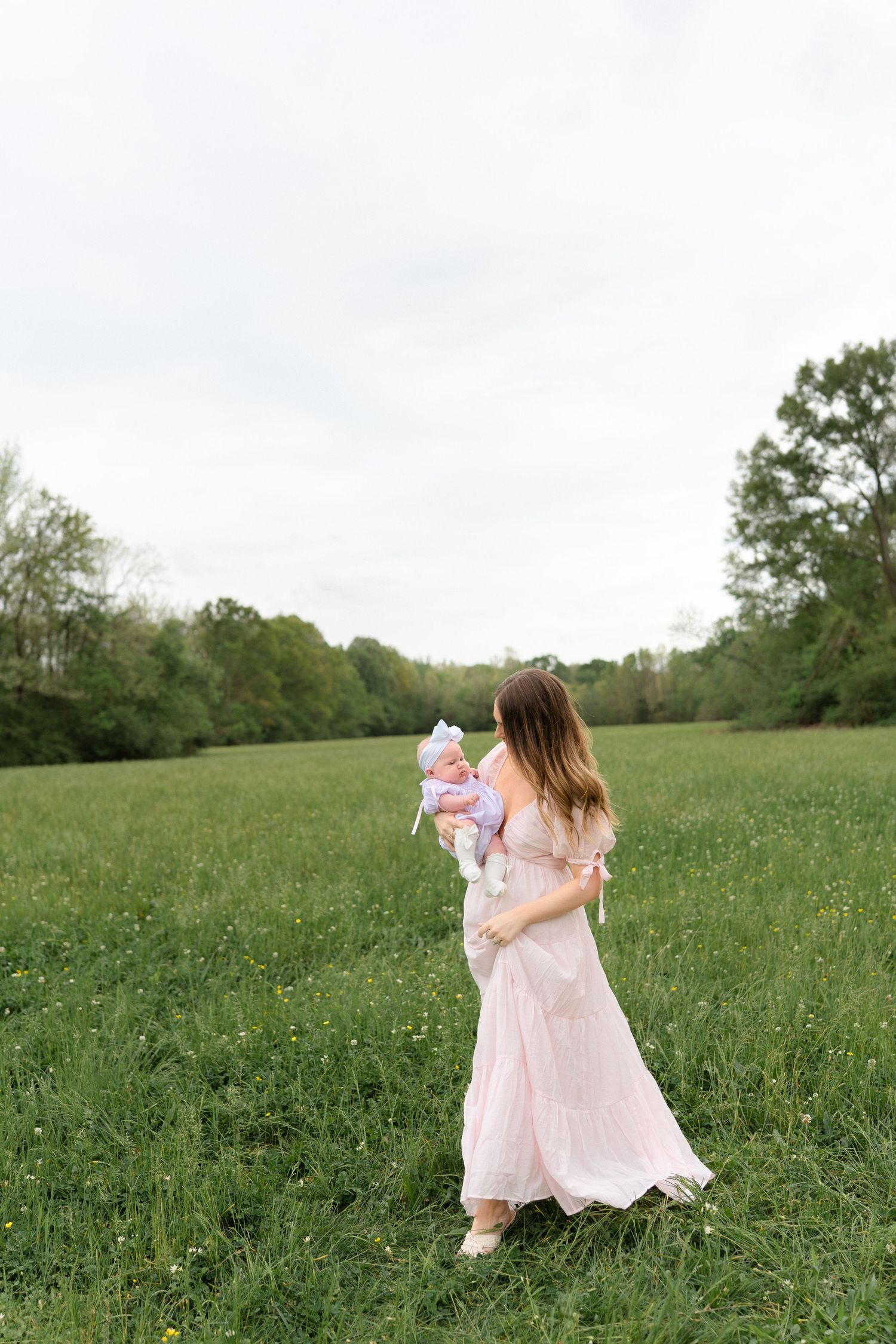A woman in a pink floral dress holds her baby girl while walking through a lush green meadow.