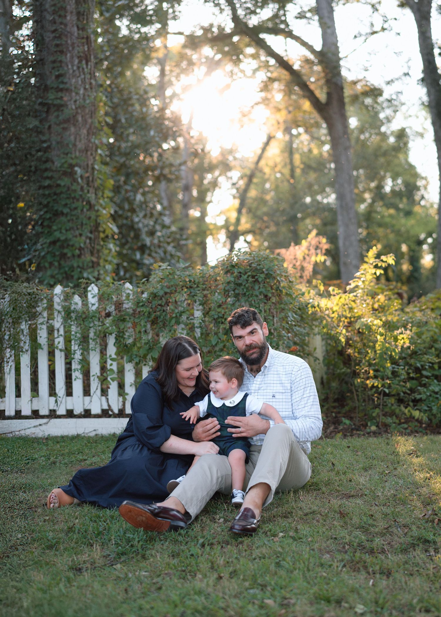 A family of three sits on grass in a sunlit garden with white picket fence and lush trees in the background.