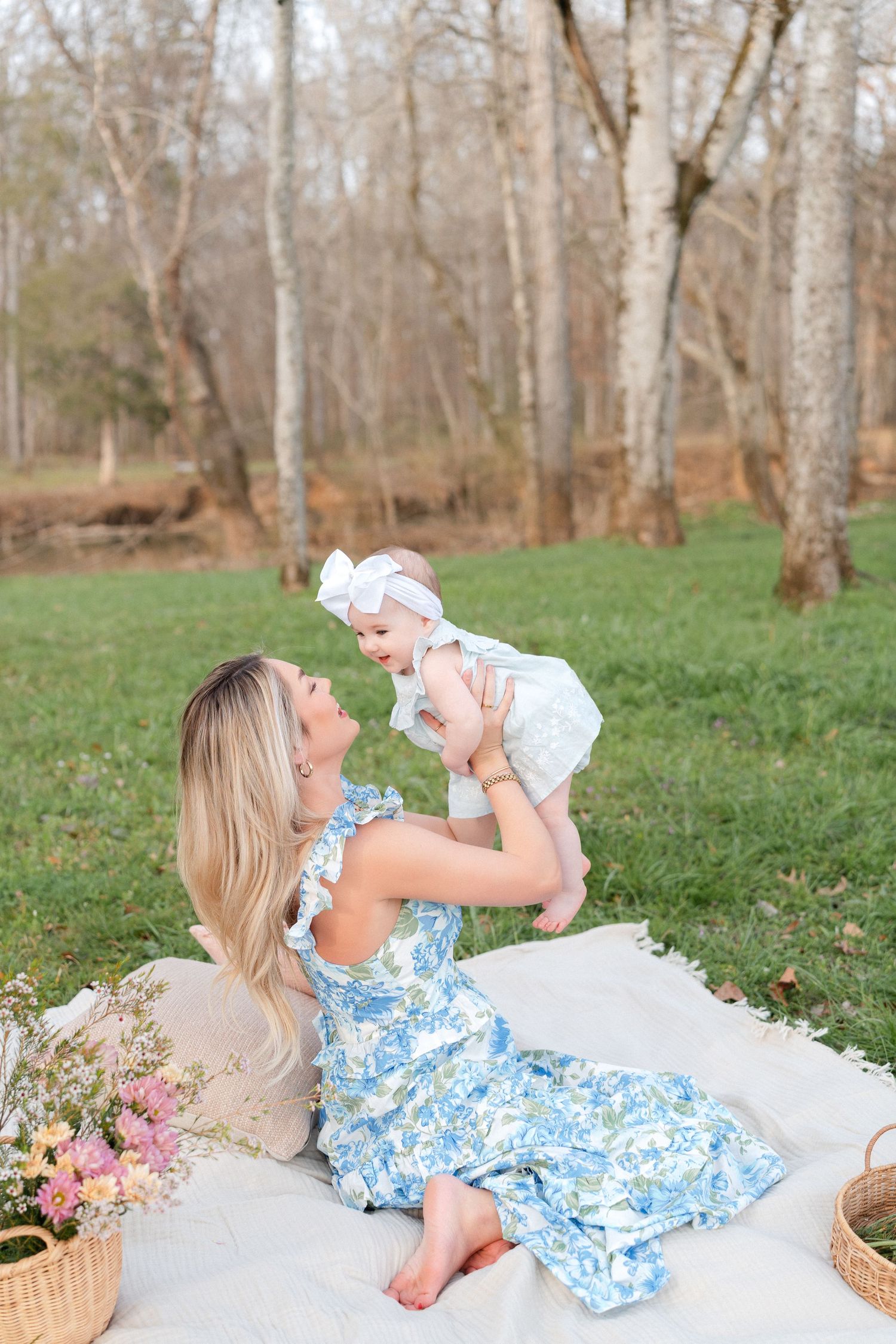 Blonde mother in blue floral dress lifts laughing baby on outdoor blanket in wooded setting.