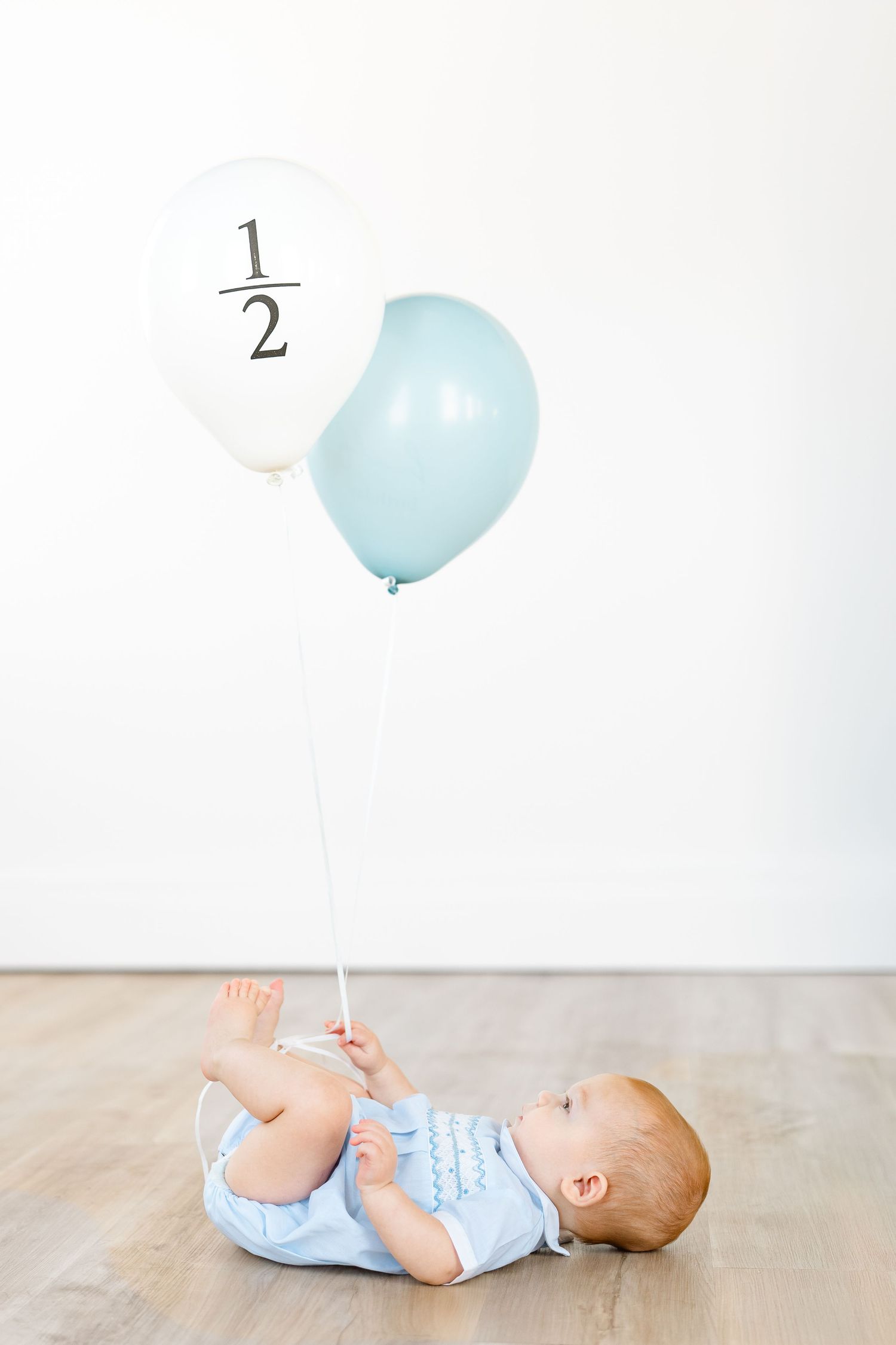 Baby boy in blue romper lying on wood floor holding white and blue balloons celebrating his half birthday.