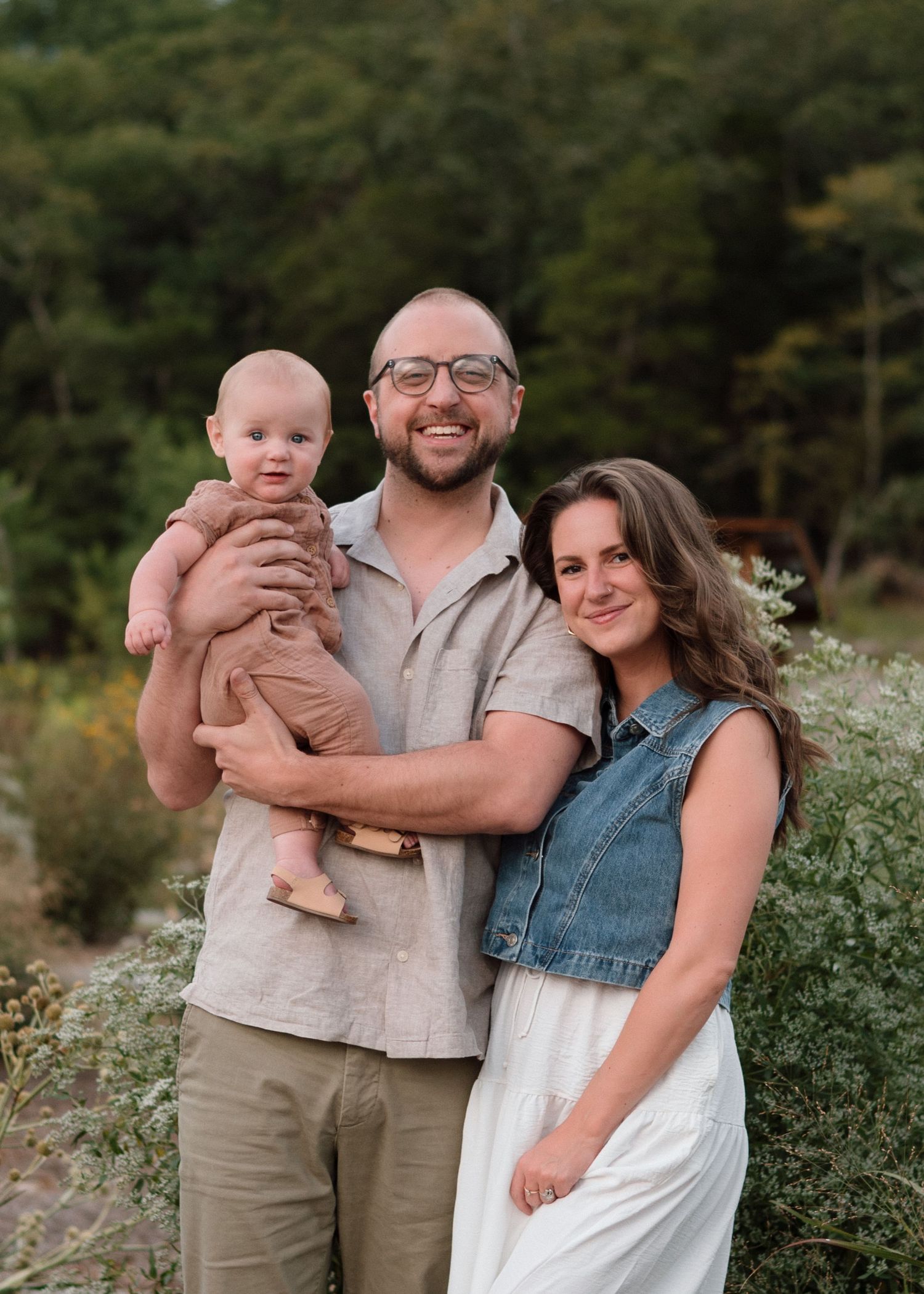 Joyful father holding baby while mother leans in, posing together in a lush green outdoor setting.