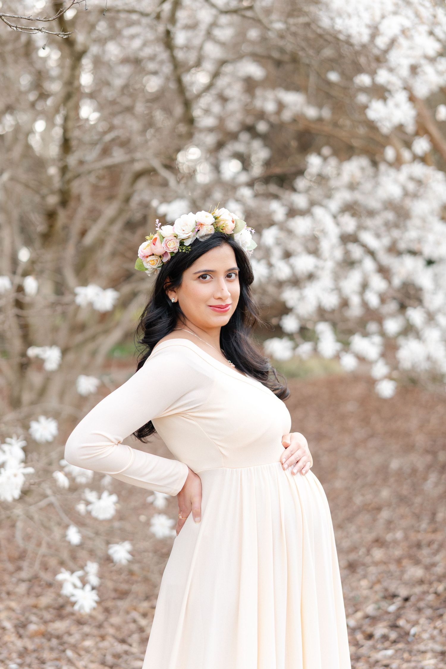 Pregnant woman in cream maternity gown and floral crown poses before white magnolia blossoms.