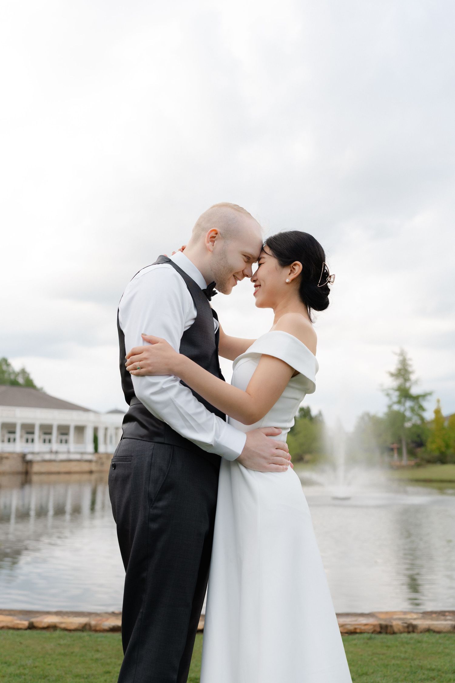 A bride and groom share a romantic embrace near a fountain and pond at an outdoor wedding venue.