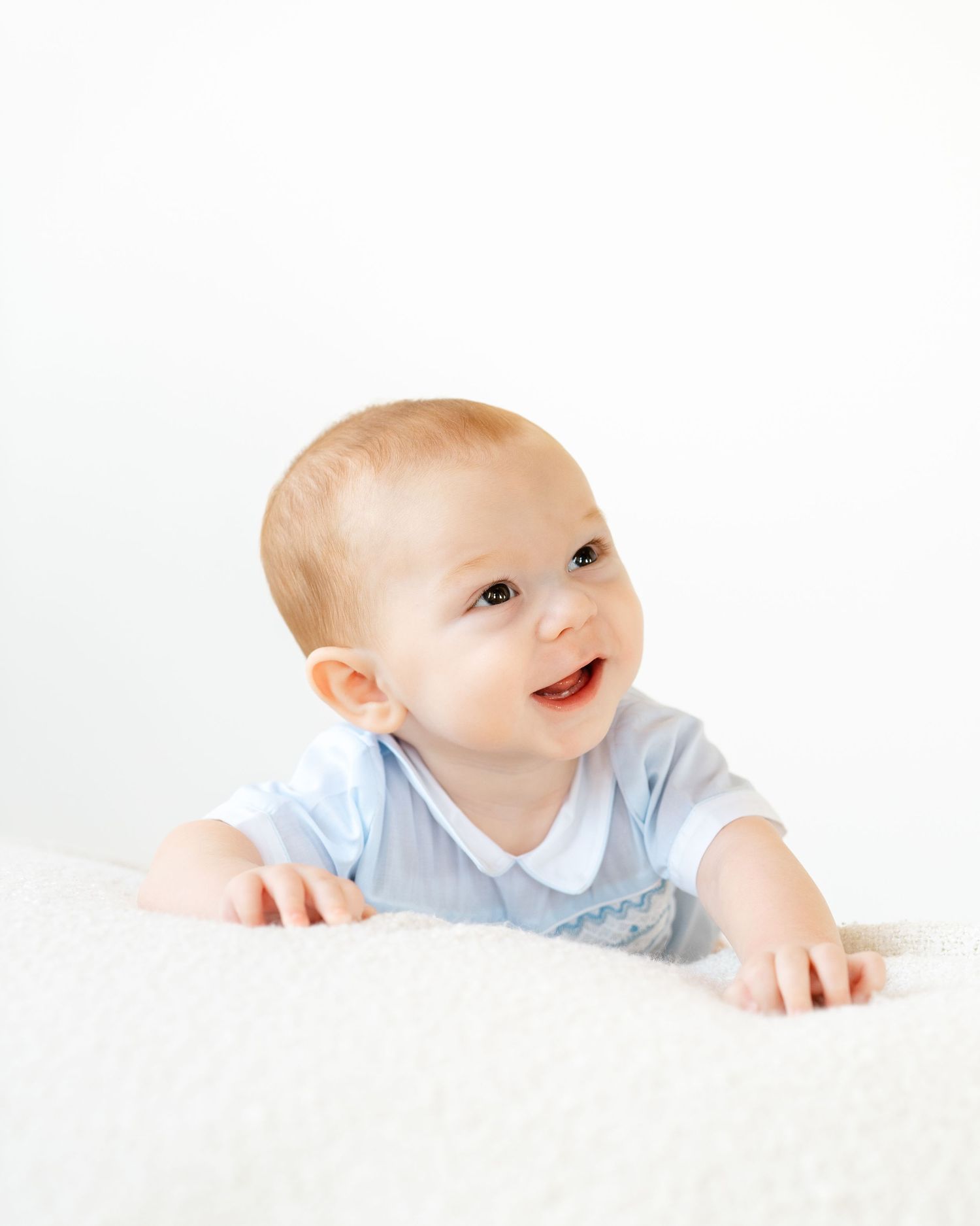 Smiling baby in blue romper doing tummy time on white fluffy blanket.