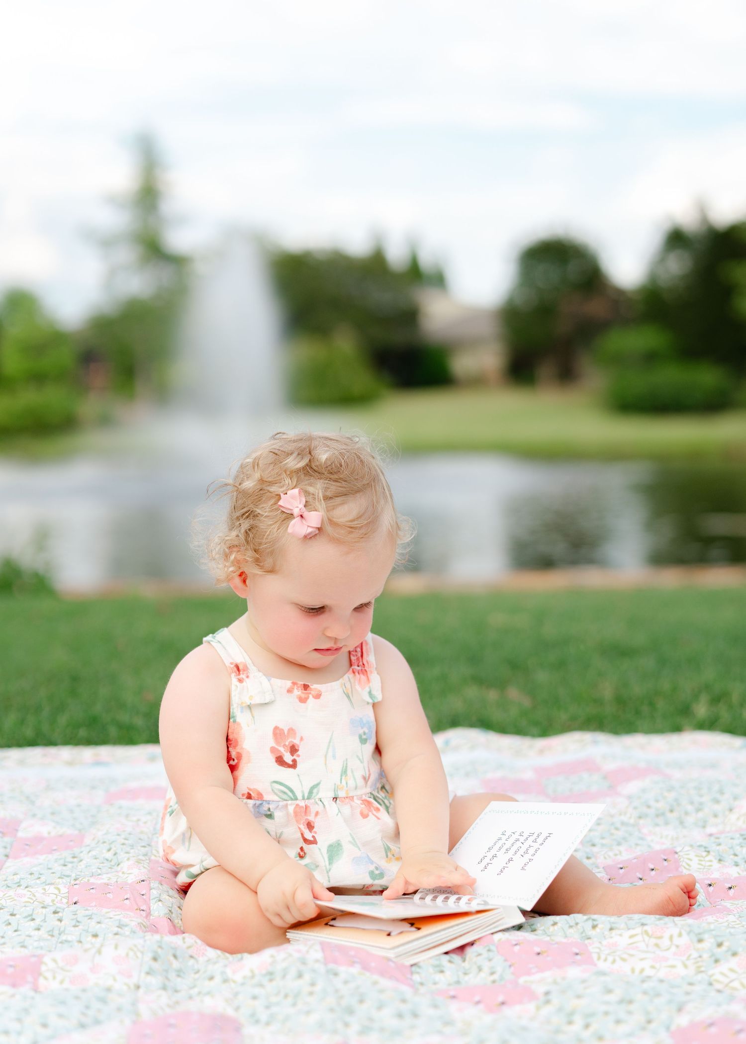 Curly-haired toddler girl in floral dress reading a book on a quilt outdoors near a fountain.