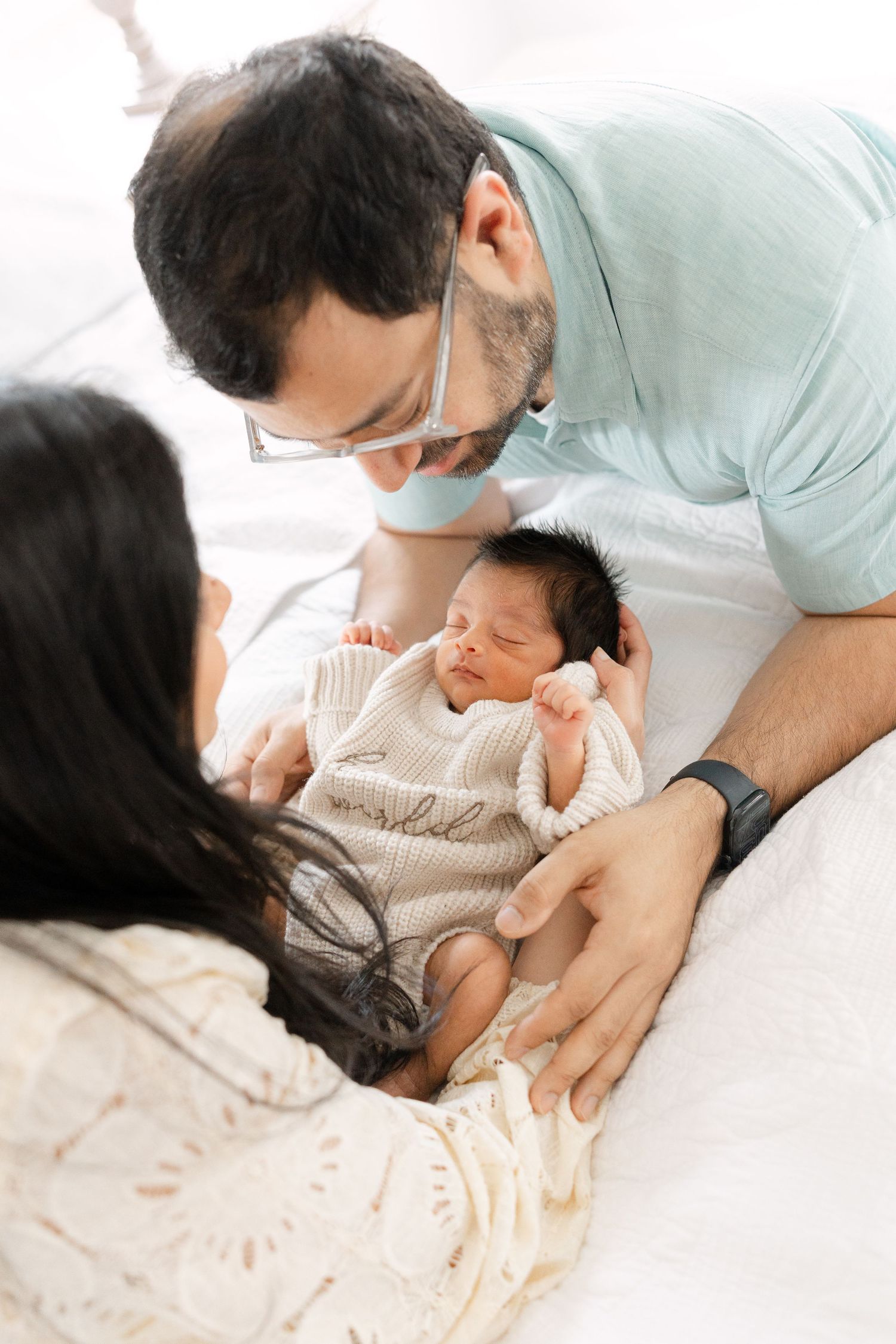 Parents in teal and white lean over bed gazing lovingly at their sleeping newborn baby.