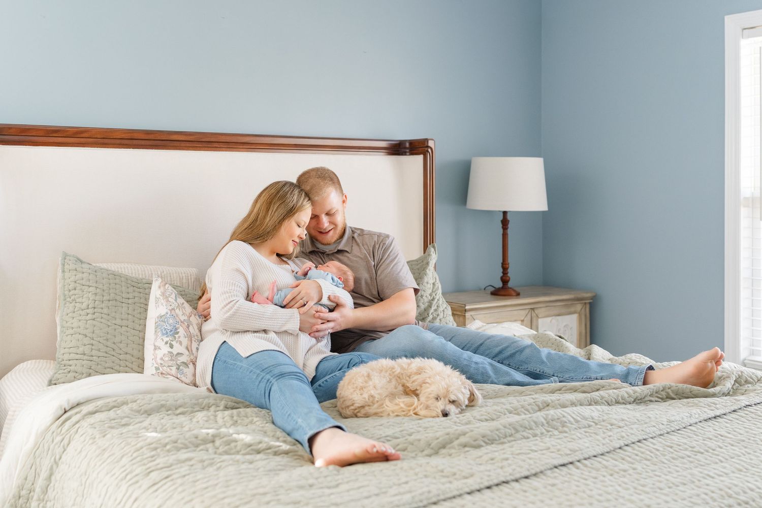 New parents sit on bed cradling newborn while fluffy white dog rests beside them.