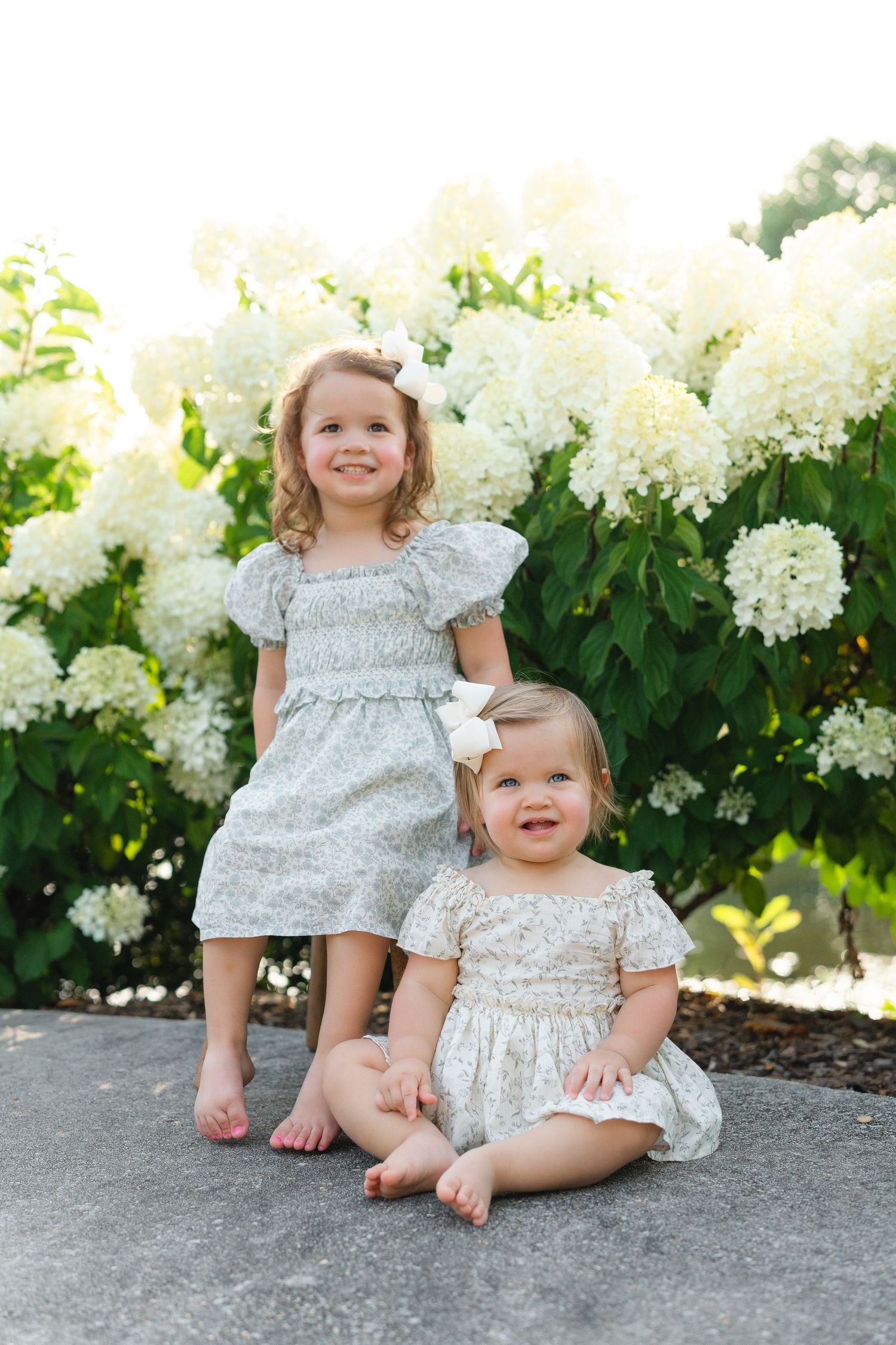 Two young sisters in floral dresses pose before white hydrangeas, one standing and one sitting on stone pavement.