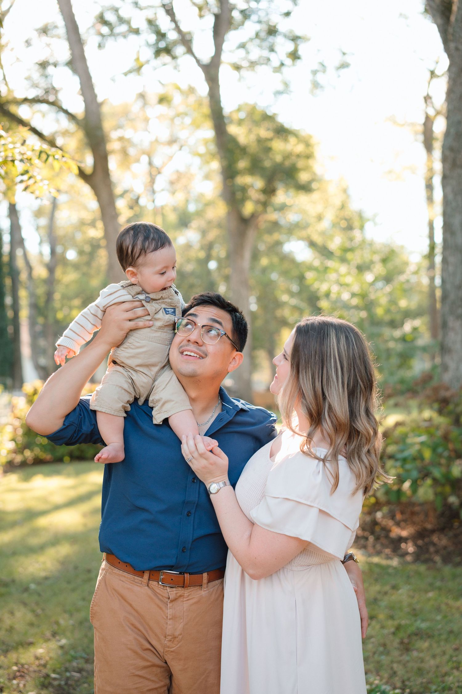 A happy young family poses outdoors, father lifting baby while mother smiles in a sunlit park setting.
