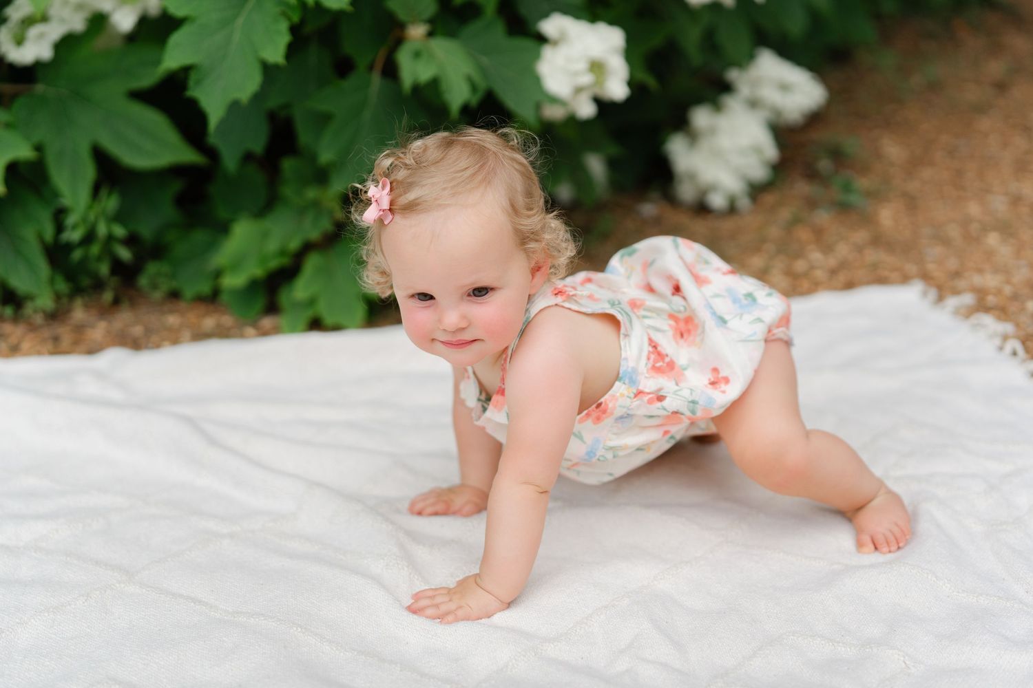 A smiling baby girl in a floral romper crawls on a white blanket outdoors near lush green bushes.