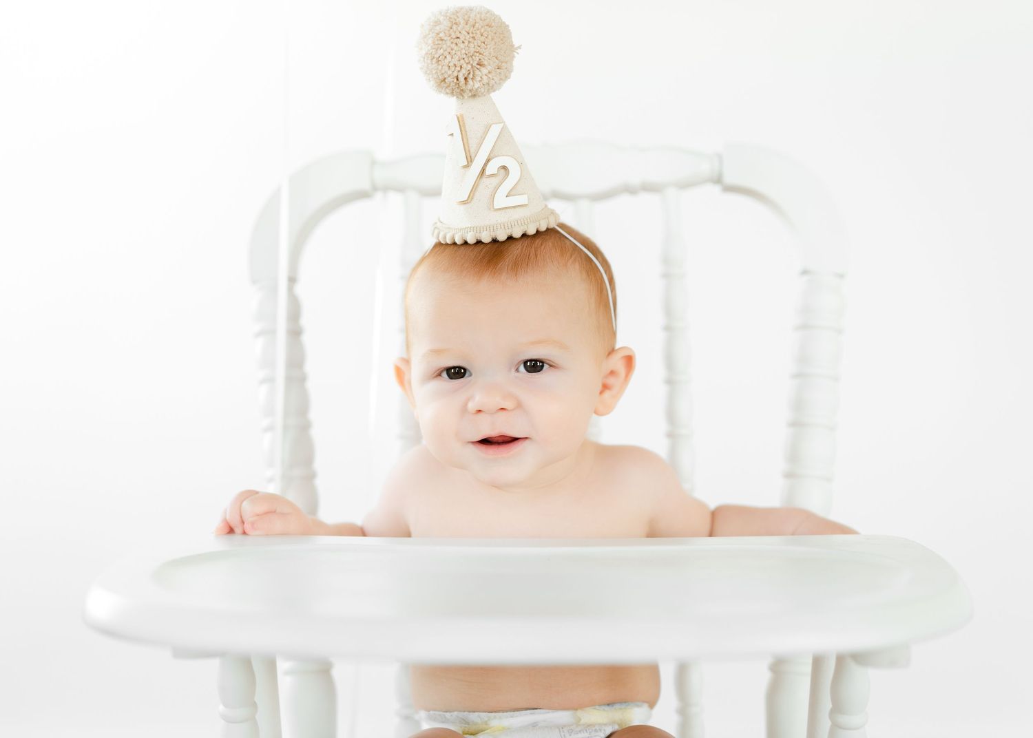 Adorable baby in white high chair wearing a half birthday party hat, celebrating their 6-month milestone.