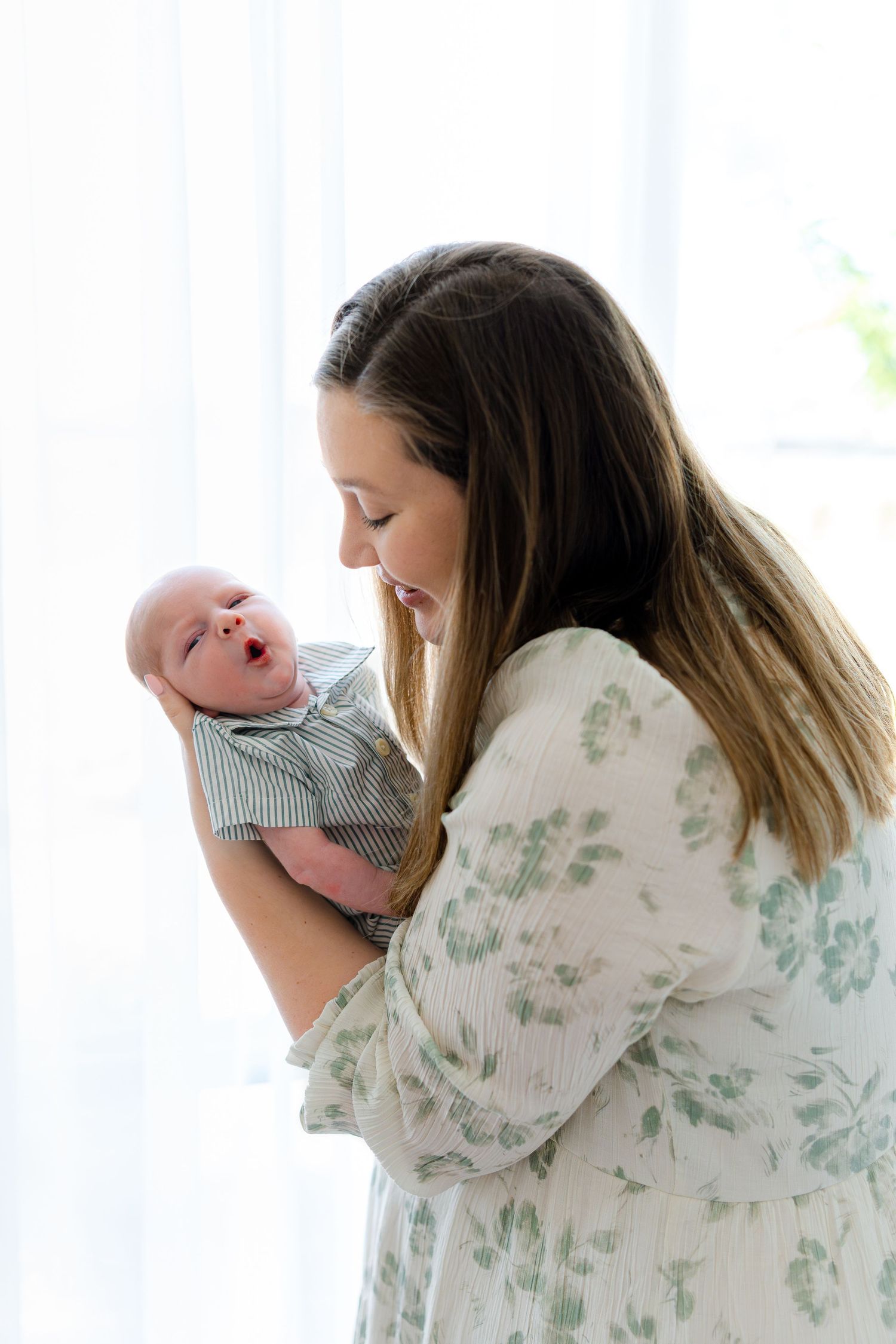 A young mother in a floral dress lovingly holds her newborn baby near a bright window.