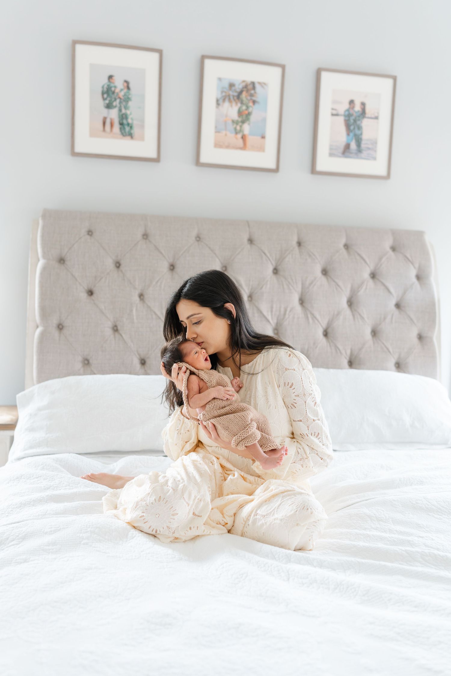 A mother in cream lace pajamas sits cross-legged on a white bed kissing her swaddled newborn baby.