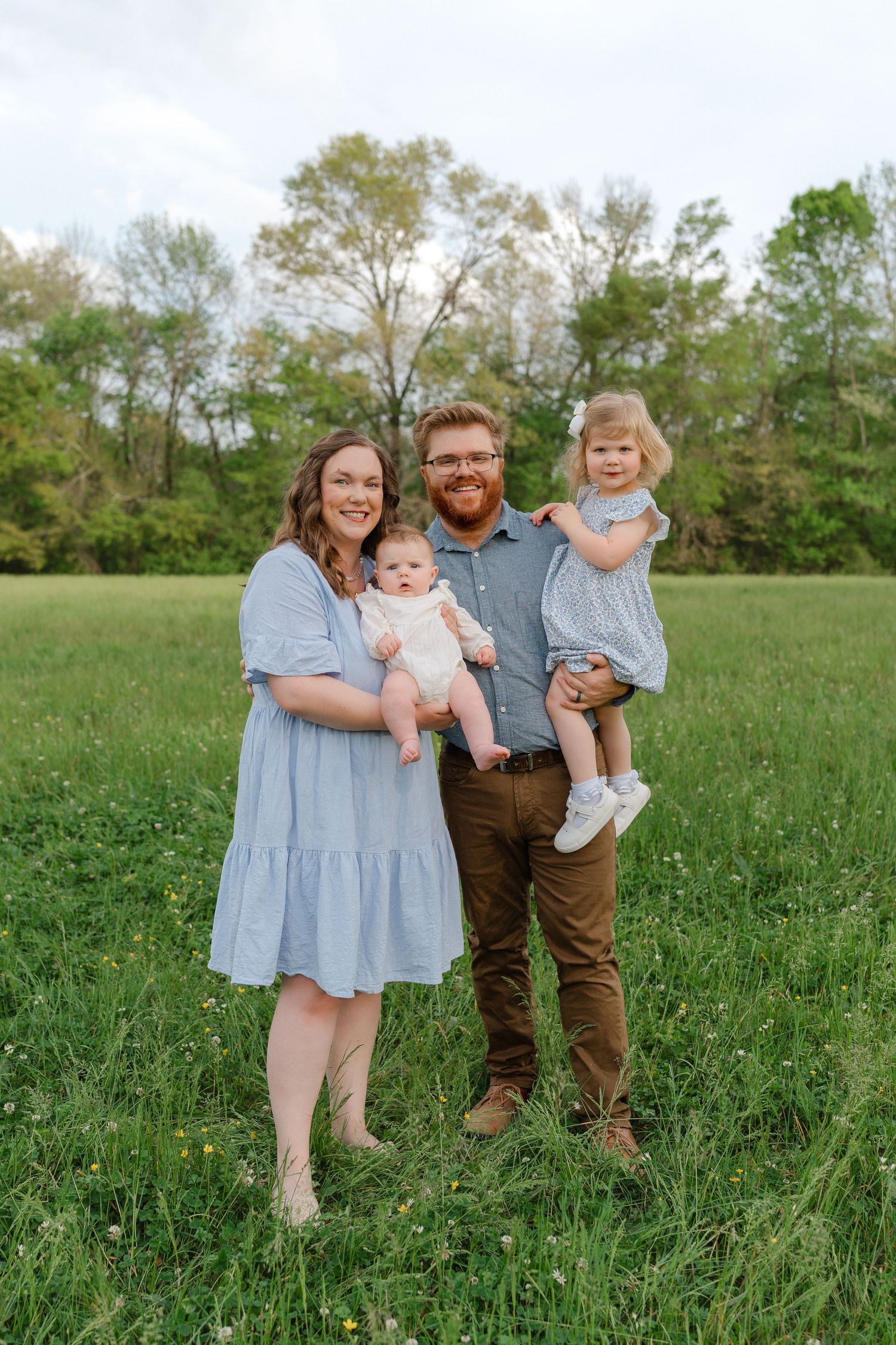 A smiling family of four stands in a green field, parents each holding a young child in coordinating blue outfits.