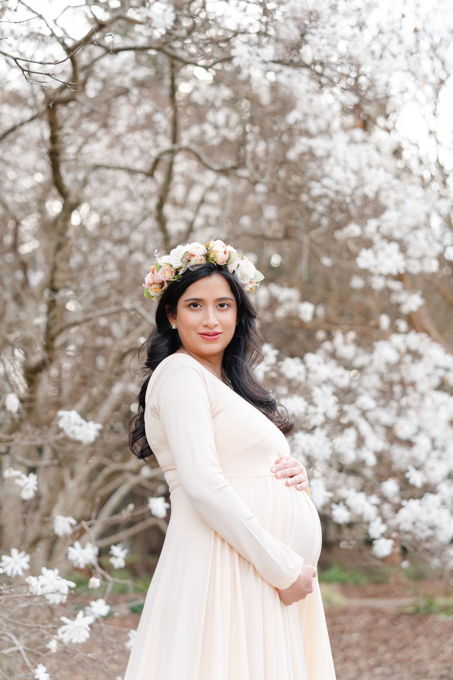Pregnant woman in cream gown and floral crown poses before blooming white magnolia tree.