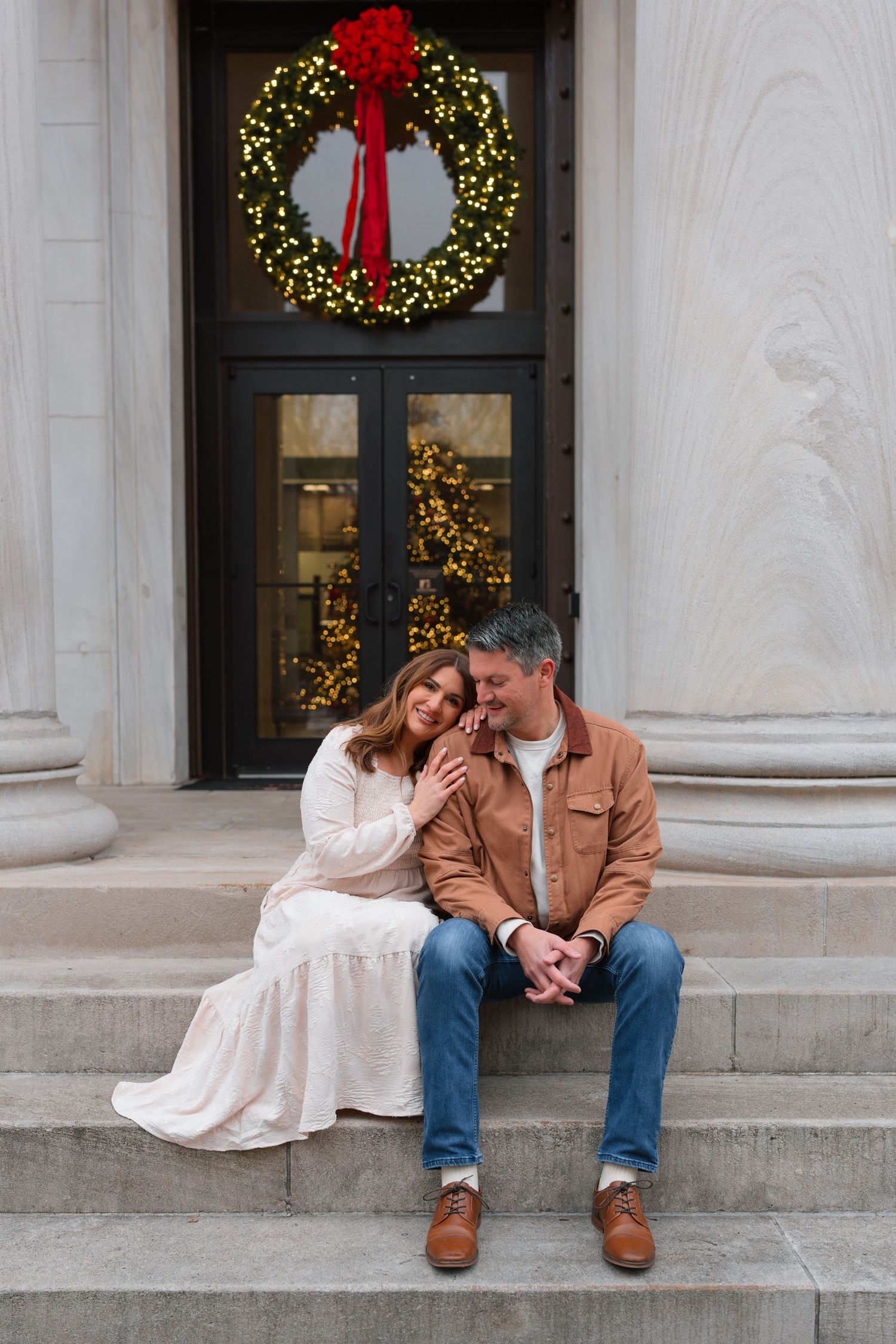 Engaged couple sits on steps beneath a glowing Christmas wreath, sharing a tender romantic moment.