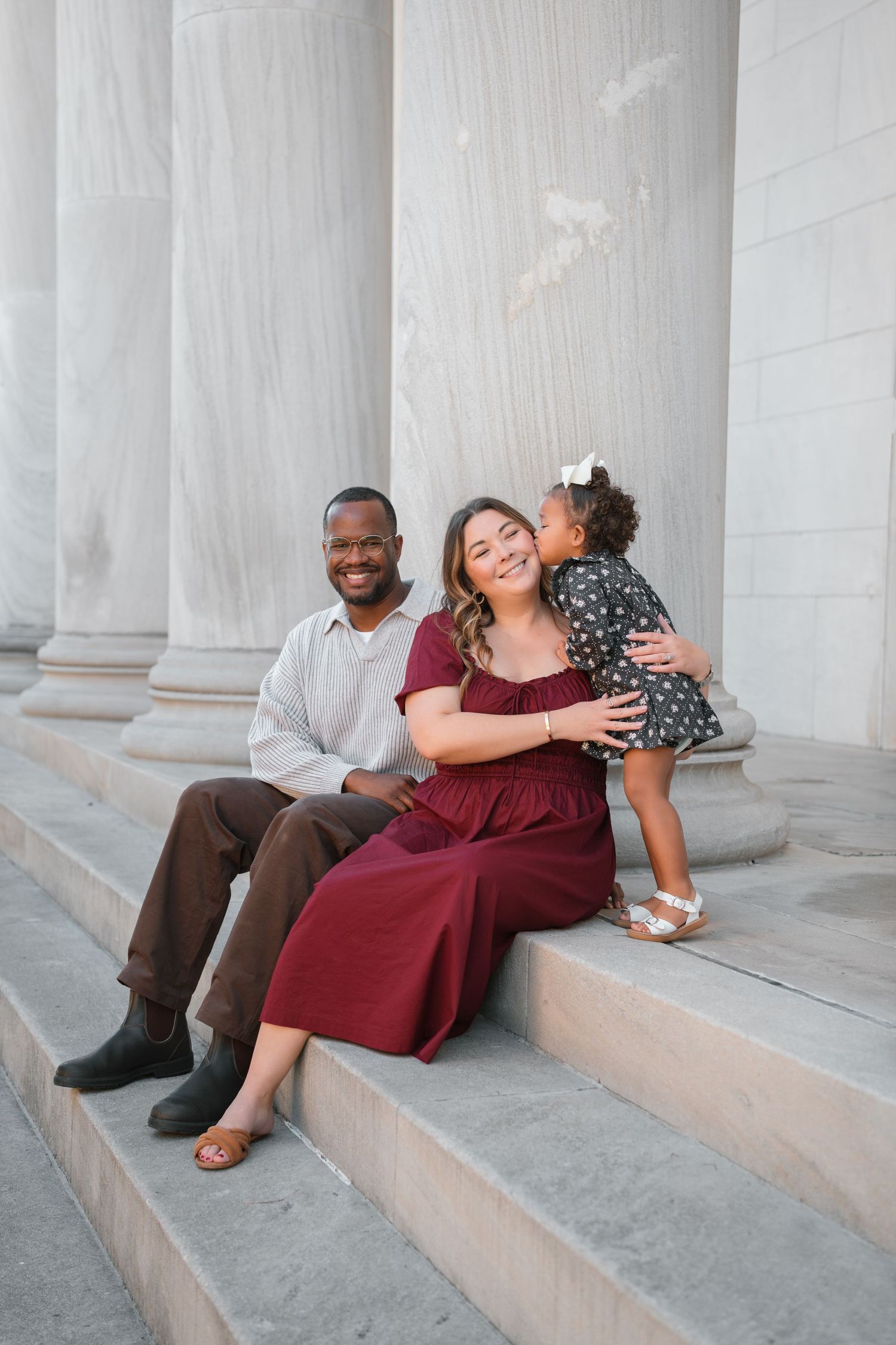 A smiling family of three sits on marble steps, with a toddler kissing her mother's cheek.