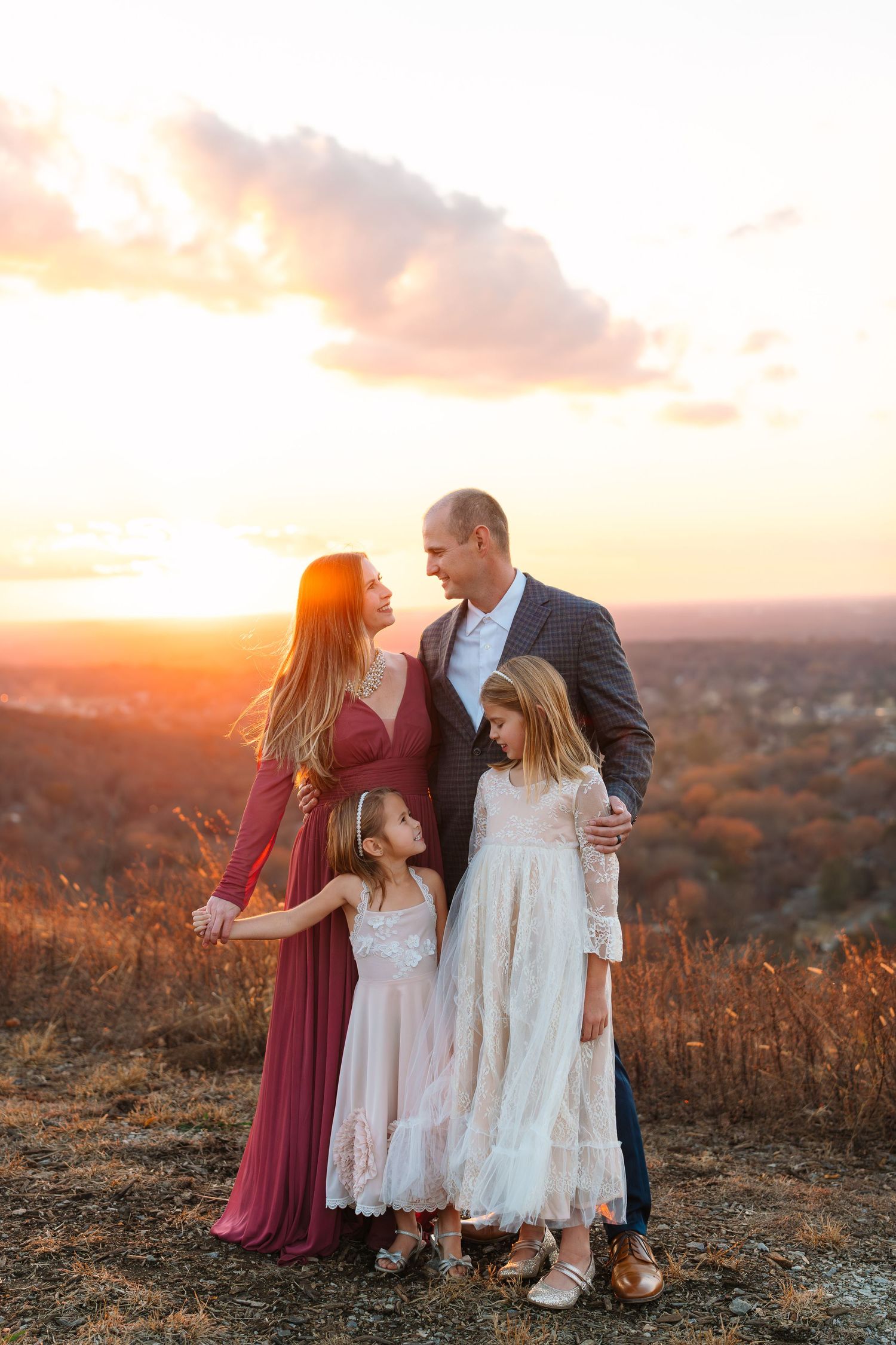 A family of four poses at golden hour sunset on a hilltop, parents embracing two daughters.