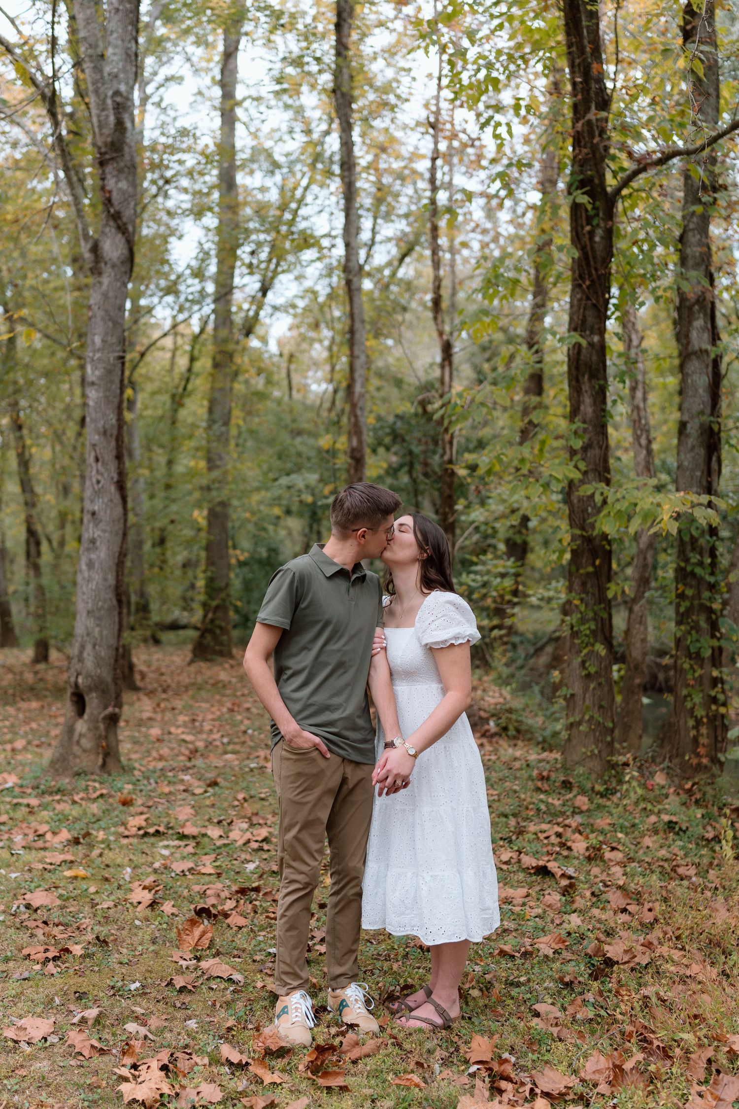 Couple sharing a kiss while holding hands in an autumn forest with fallen leaves.