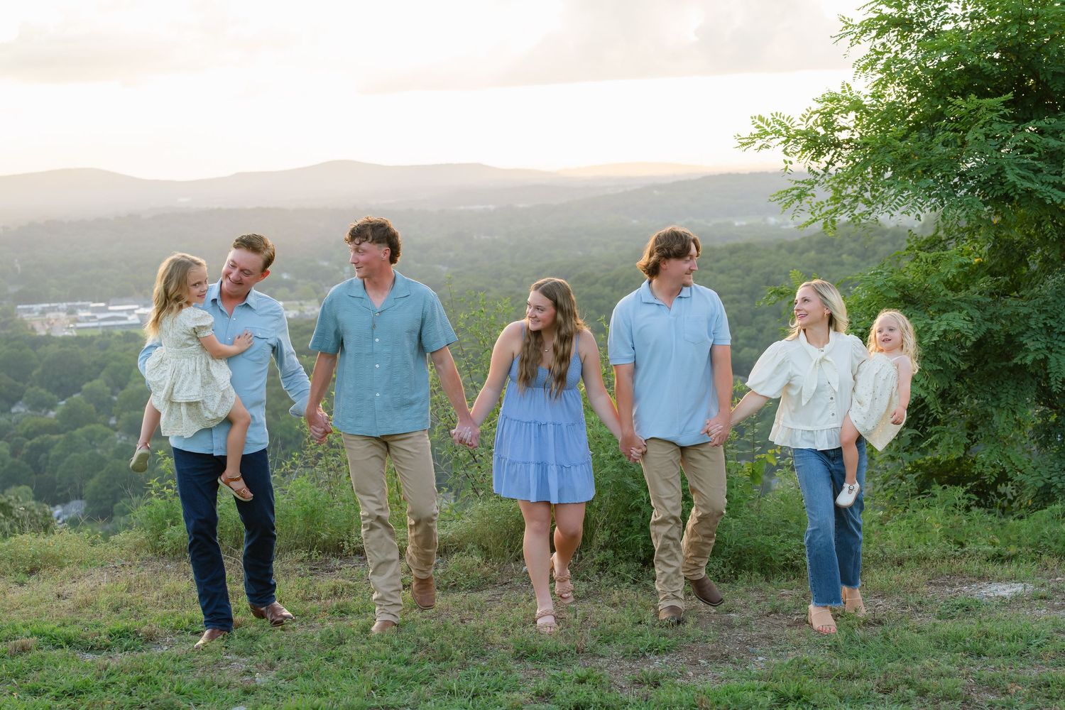 Family of six holding hands walking on a grassy hilltop with scenic mountain views at sunset.