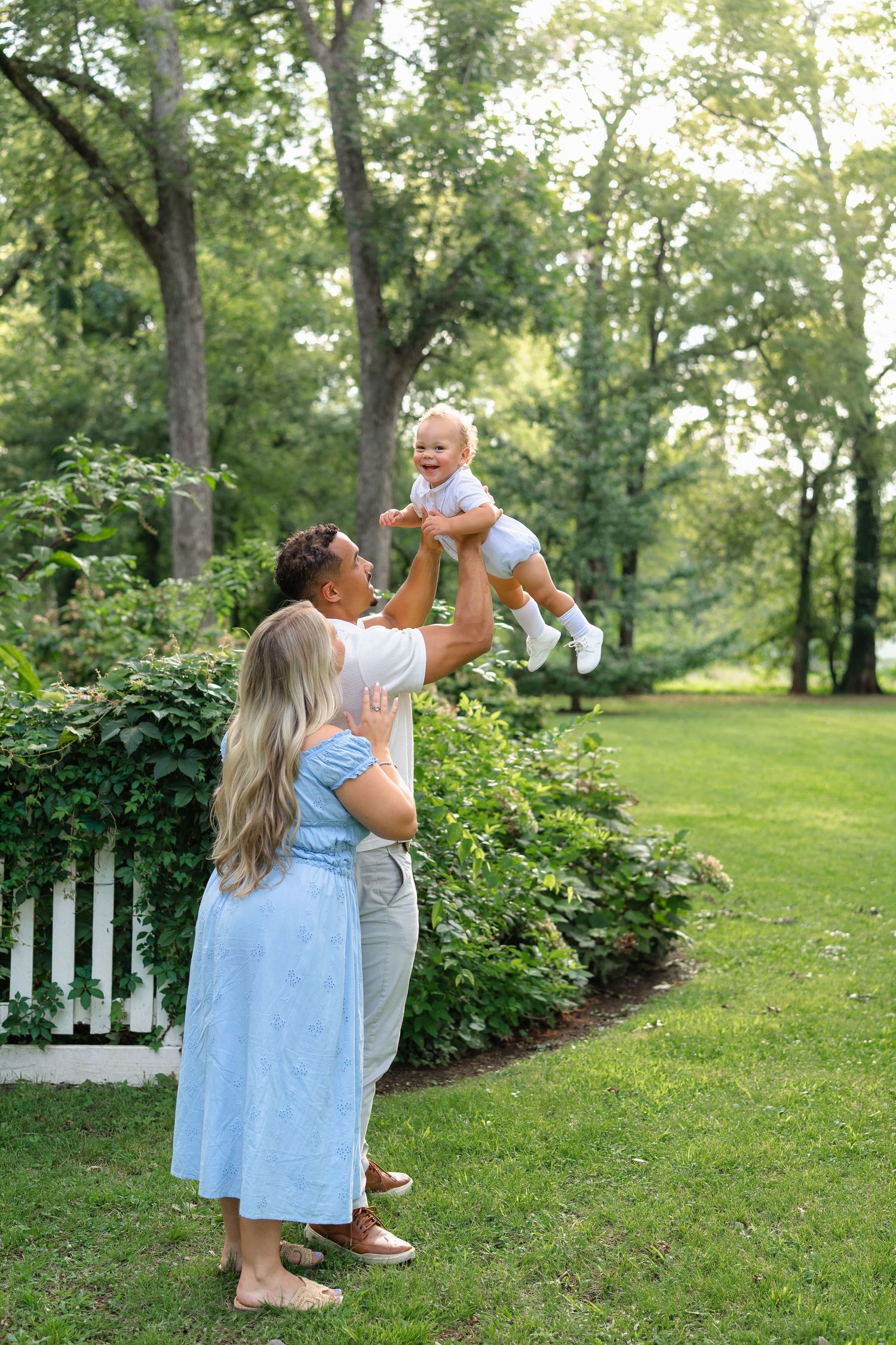A couple plays with their baby in a lush green backyard, lifting the laughing infant in the air.