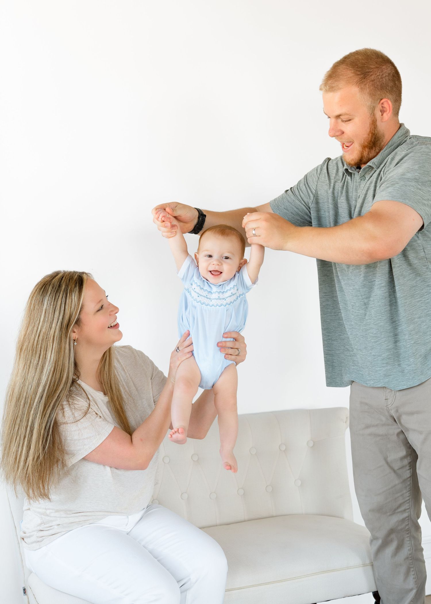 Happy family with mom, dad, and smiling baby in blue romper playing together in bright white room.